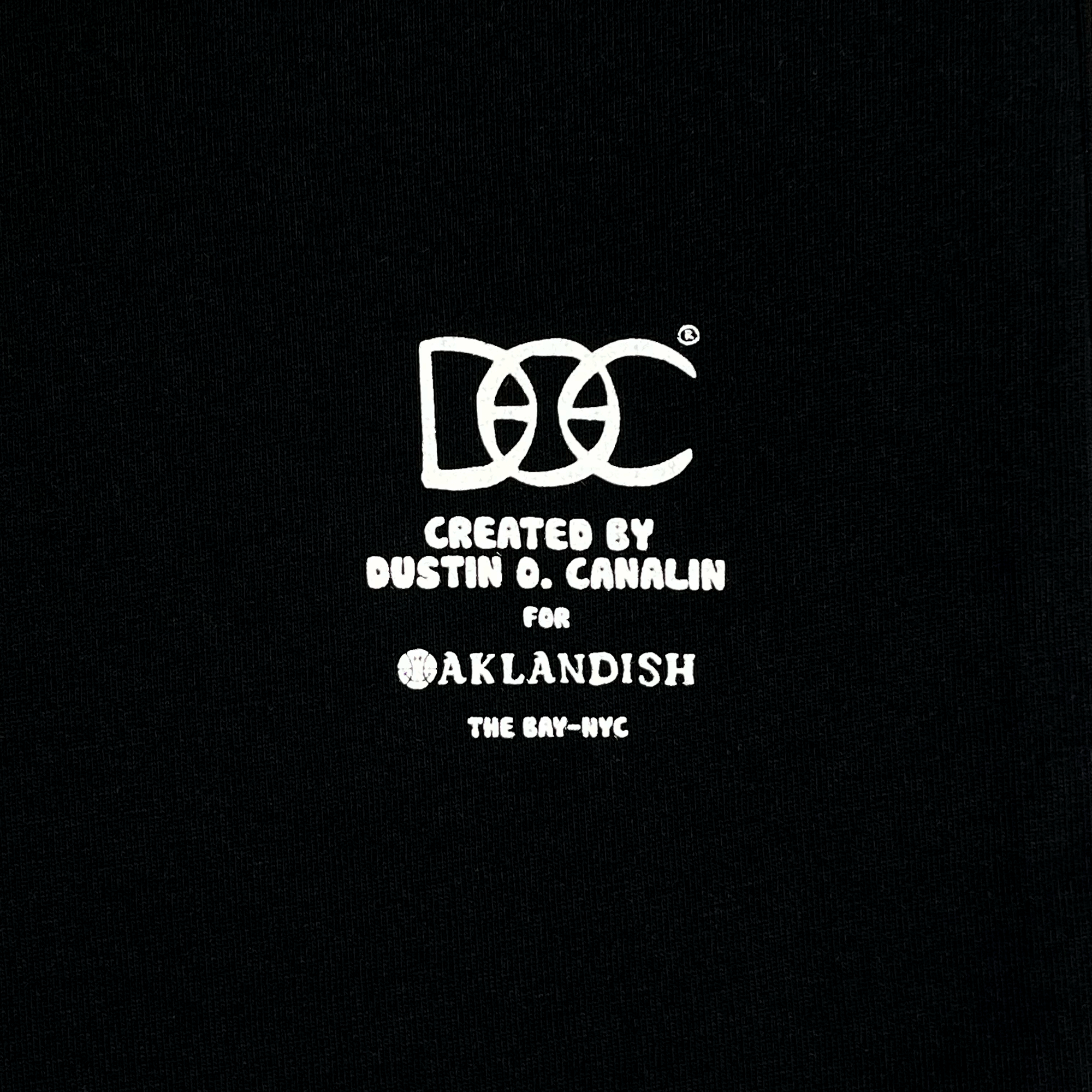 Close-up of white DOC logo Created by Dustin O. Canalin for Oaklandish The Bay-NYC wordmark on a black t-shirt.