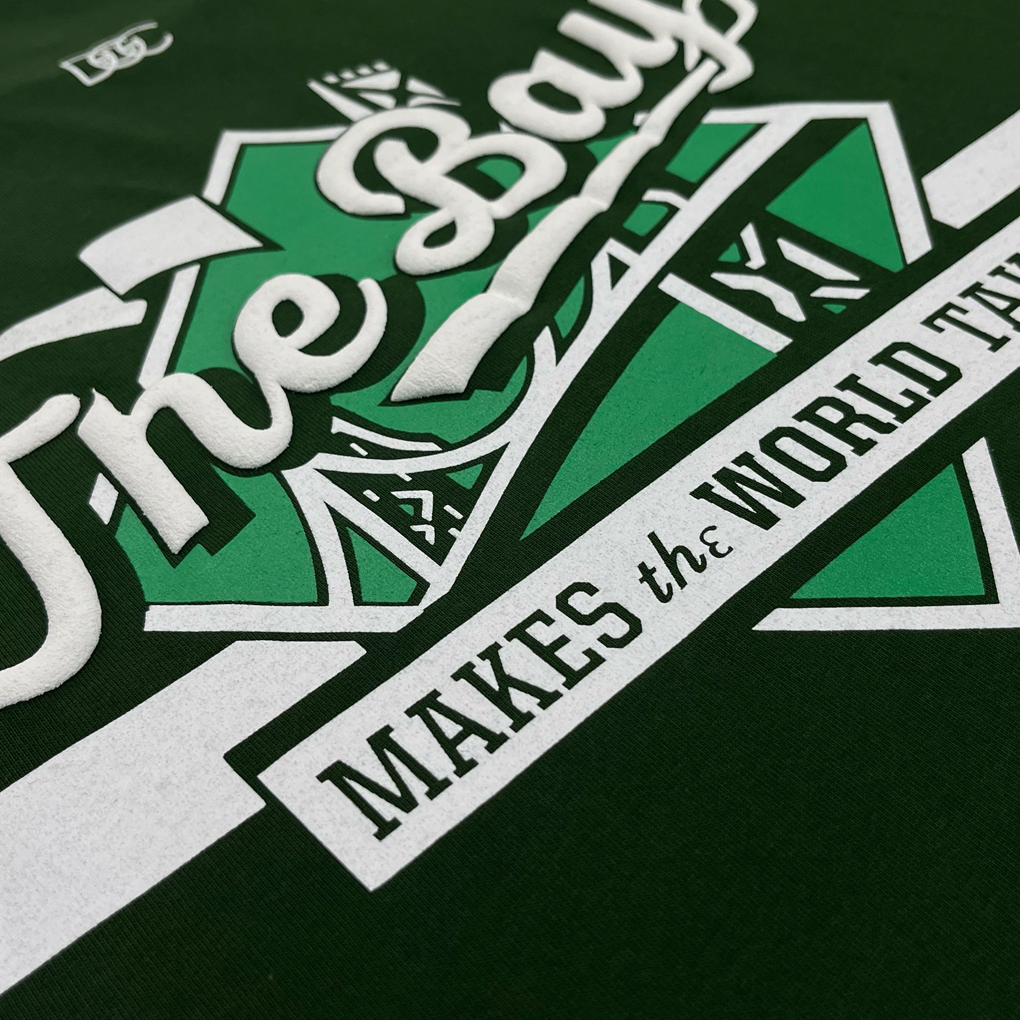 Angled close-up of a white The Bay Makes The World Takes logo graphic on the left chest wearside of a forest green t-shirt.