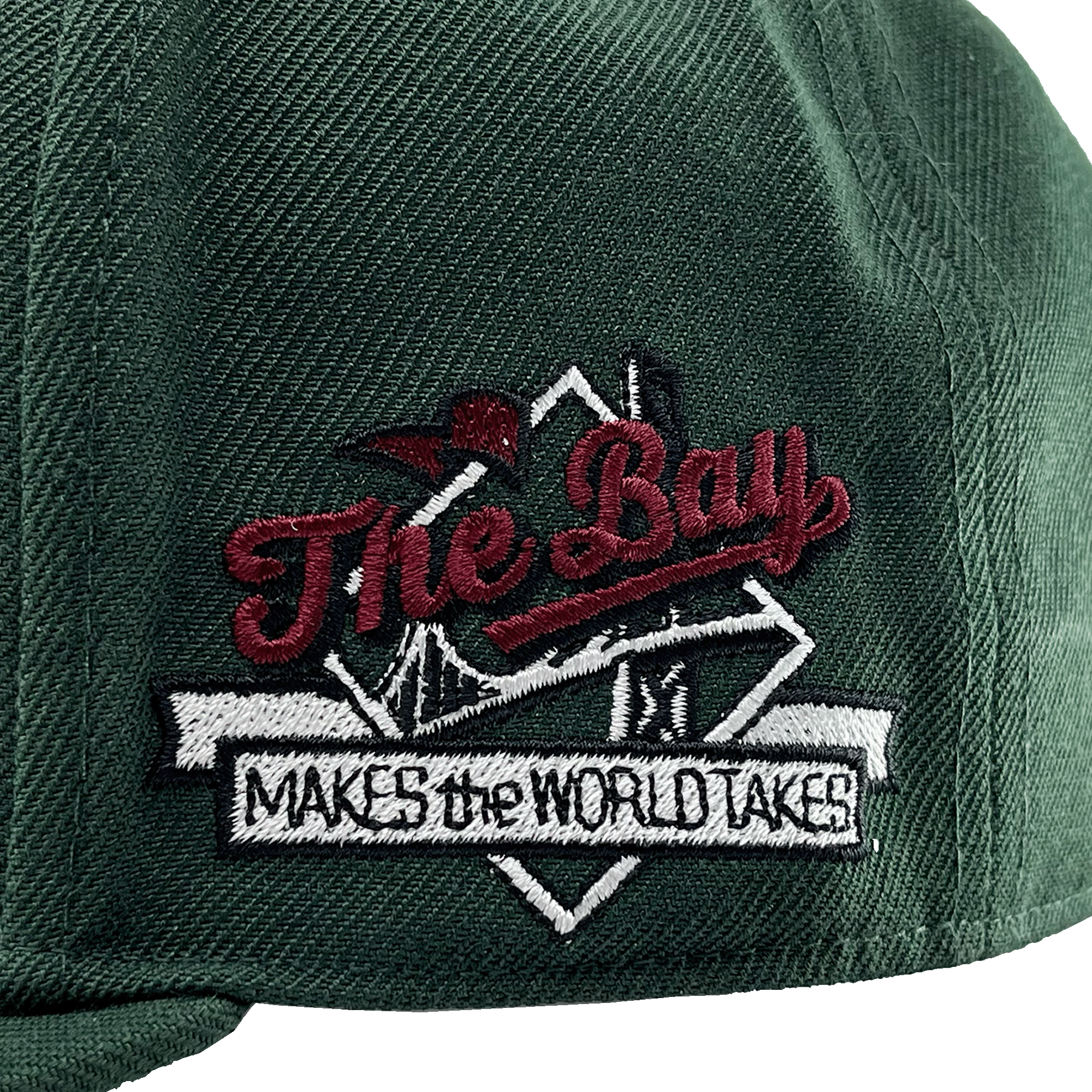 Detailed close-up of The Bay Makes, The World Takes detail on the wearer's left side of a green hat.