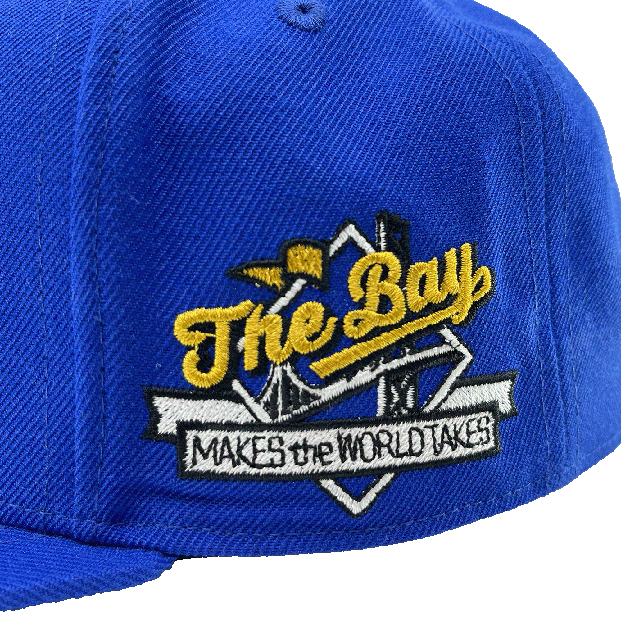 Close-up of full-colored The Bay Makes, The World Takes detail on the wearer’s left side of a royal blue hat. 