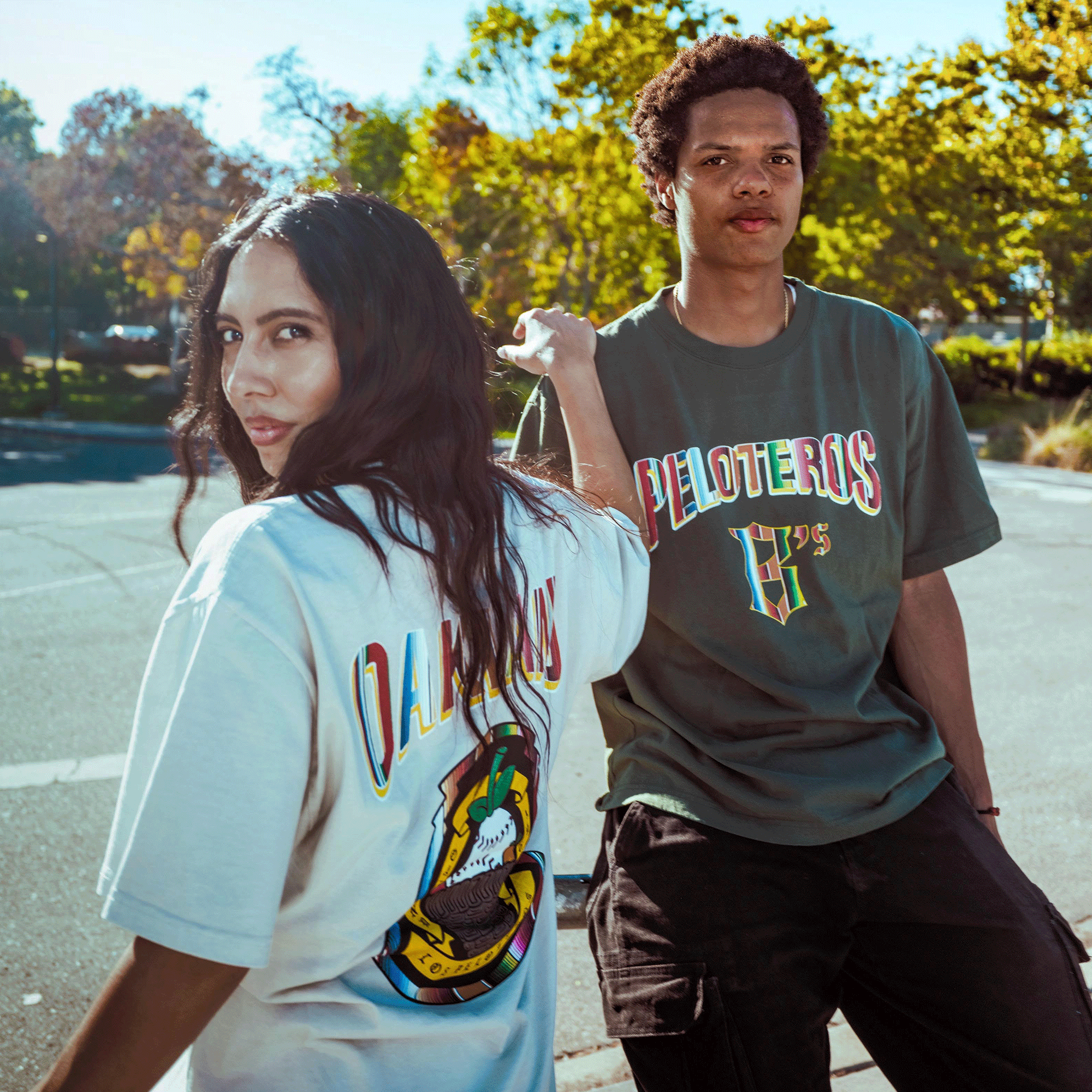 Two young adults pose outside under blue sky and trees. She wears a white tee with a colorful graphic; he sports the Oakland Ballers "Oakland Beisbol Tee," repping Oakland’s Pioneer League baseball team.