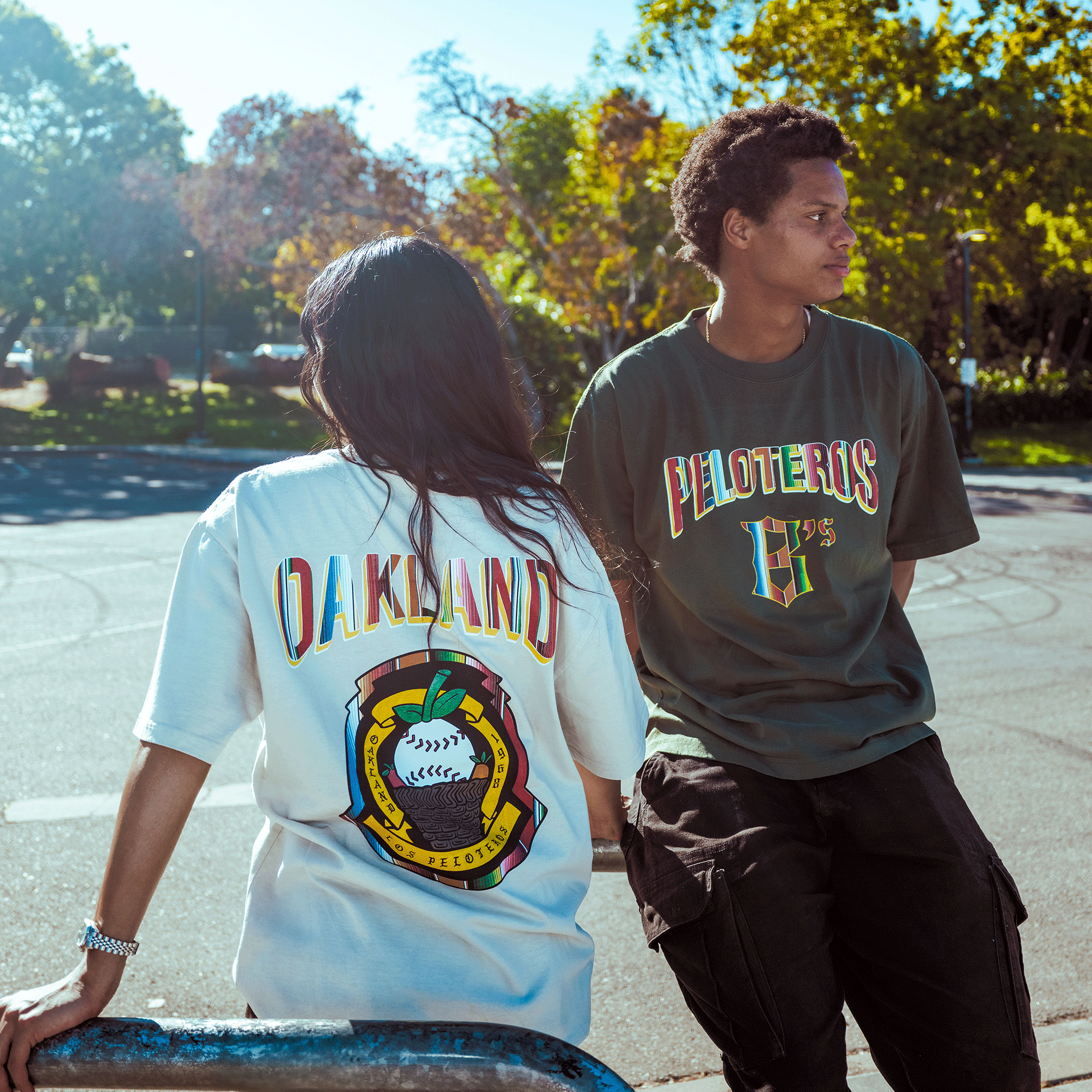 Two people stand outdoors in front of lush trees. One wears a white "Oakland Beisbol Heavy Tee" by Oakland Ballers with a baseball graphic, while the other sports a dark "Peloteros" shirt, capturing the vibrant Pioneer League spirit.