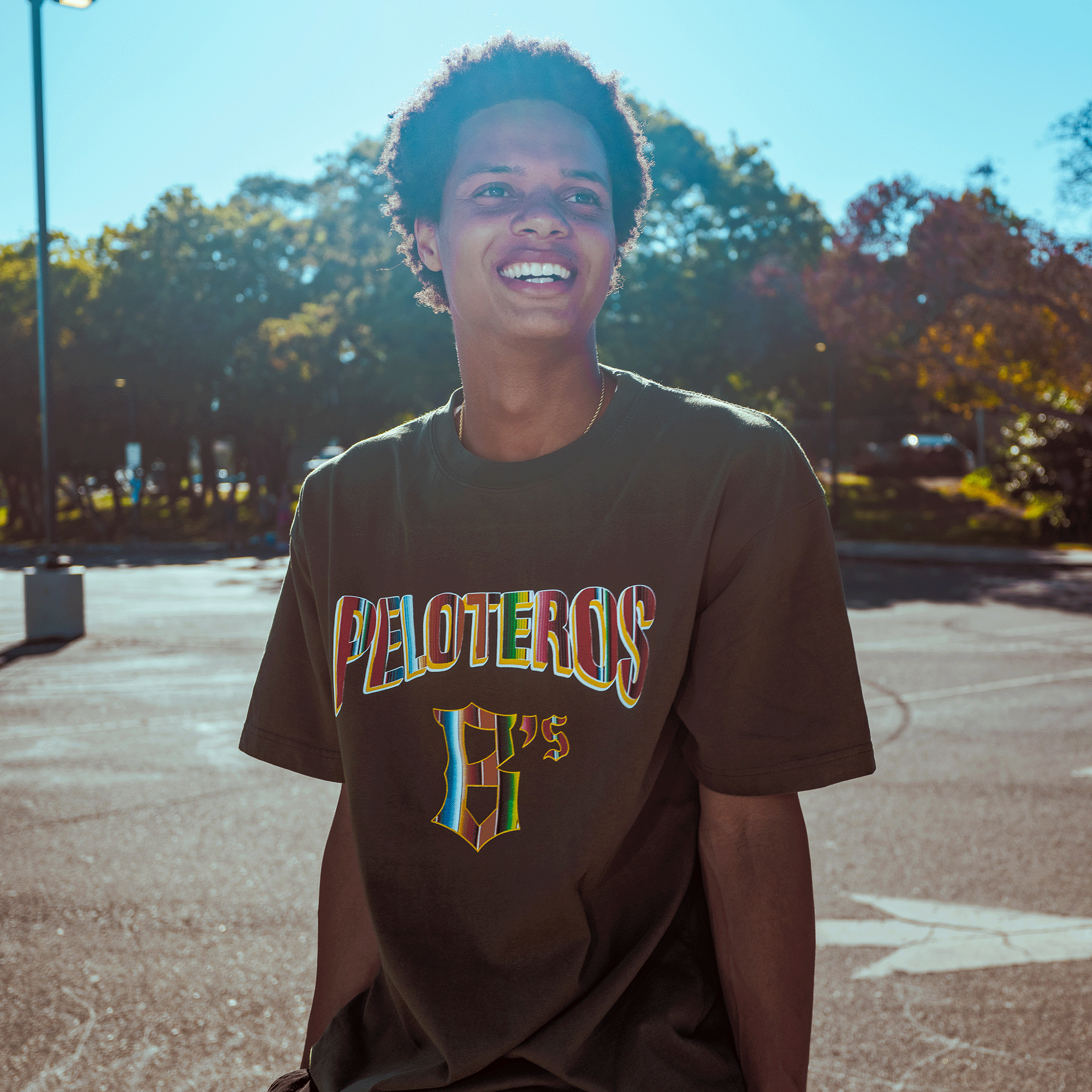 A smiling young person with short curly hair wears the Oakland Ballers Oakland Beisbol Tee outdoors, with autumn trees and a sunny sky behind them, reflecting the energetic spirit of the Pioneer League baseball team.