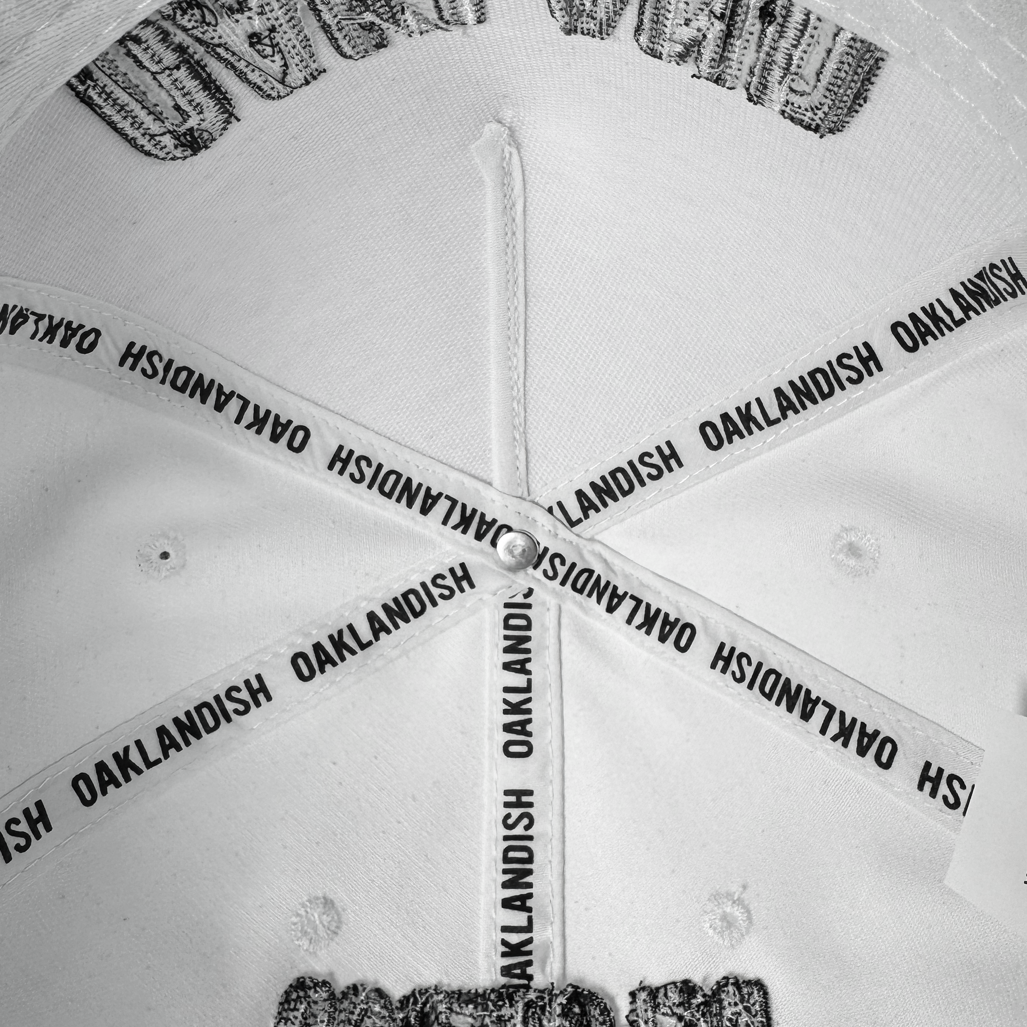 Close-up of the inside top of the white Oakland Beisbol 5 Panel Cap by Oakland Ballers. Four intersecting fabric strips, each printed with "OAKLANDISH" in black, plus white stitching and grommets evoke classic baseball team style.