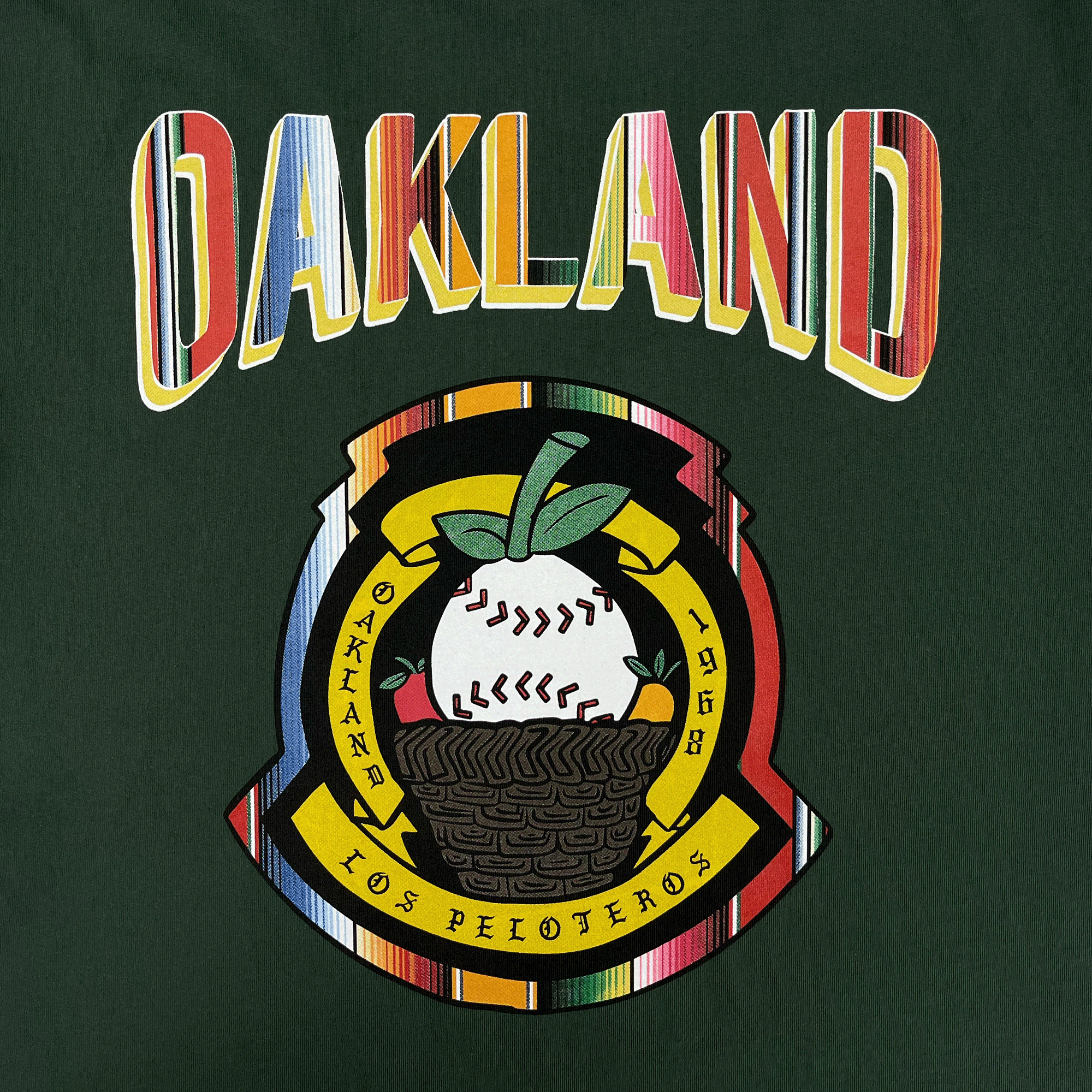 The Oakland Beisbol Tee by Oakland Ballers is a green shirt with "OAKLAND" in bold, colorful letters above a circular patch featuring a baseball plant in a basket and the text "OAKLAND - 1968 - LOS PELOTEROS.