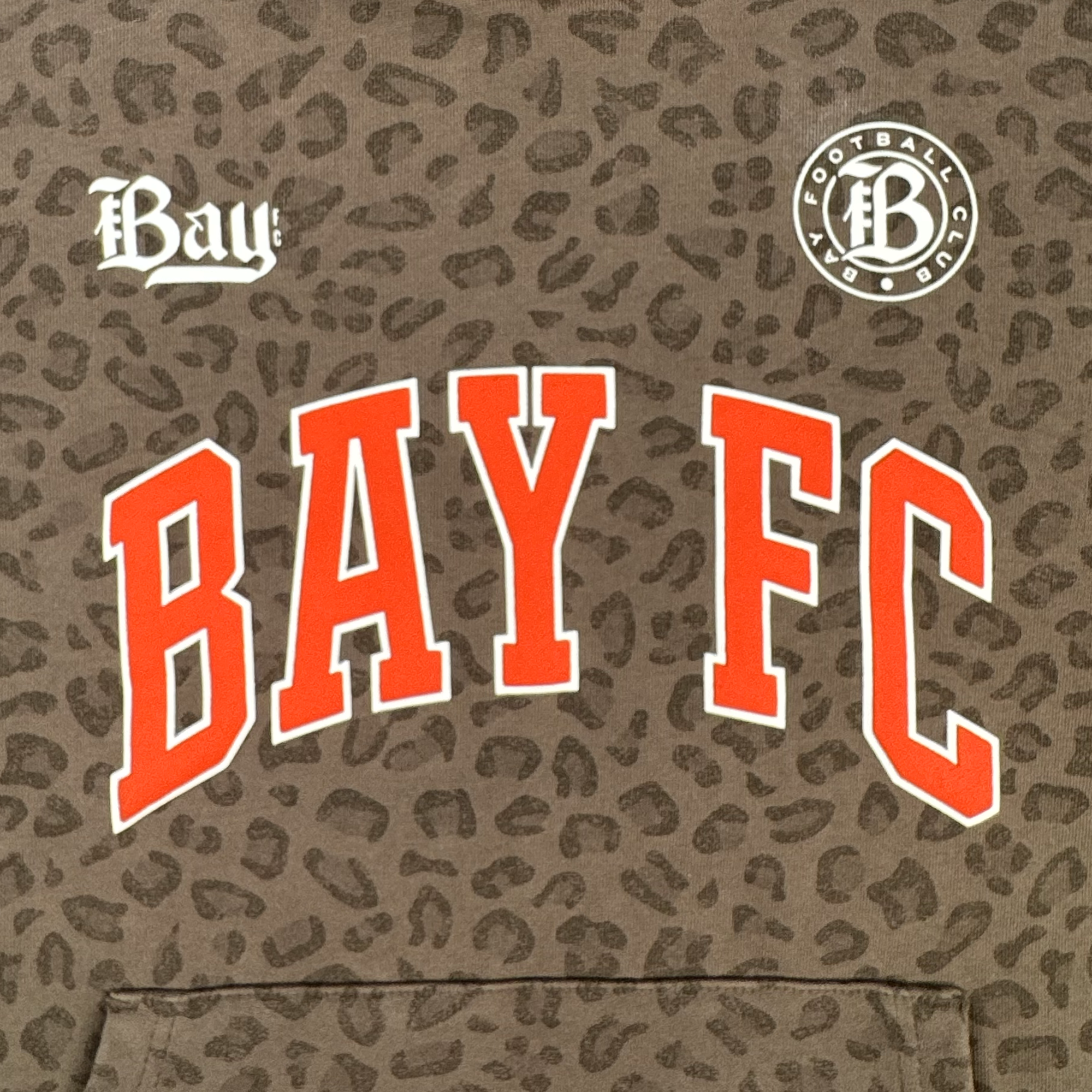 The Bay FC Leopard Hoodie by Bay FC features a brown leopard camo print, "BAY FC" in bold red and white on the chest, "Bay" in white on the left, a circular Football Club logo on the right, and a front pouch pocket.