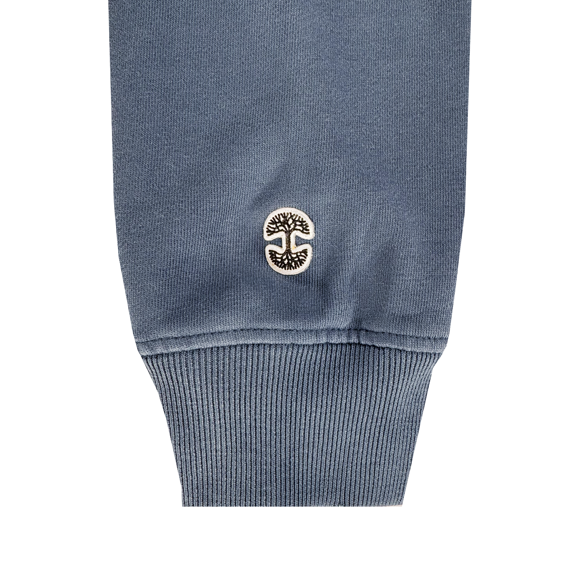 A close-up of the Oaklandish Homage Hoodie’s blue ribbed cuff, made from 100% cotton, shows a small embroidered black and white circular patch with a stylized tree logo near the hem.