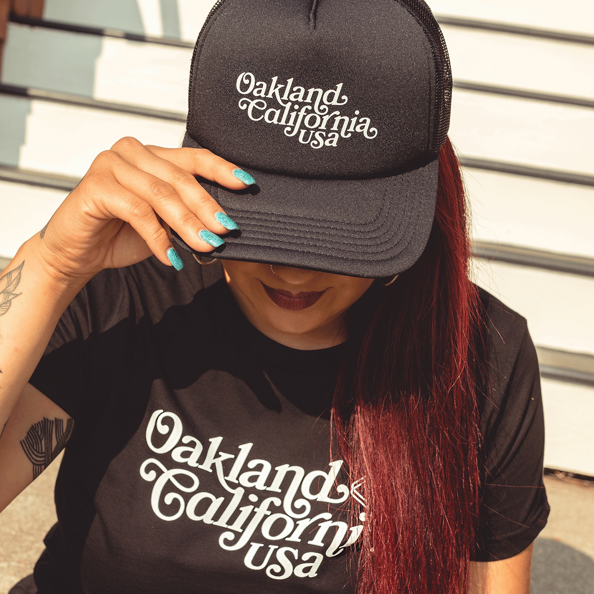 Model wearing a black trucker cap tilted forwardwith foam front panel, mesh back, adjustable back, and white Oakland, California, USA wordmark on the crown.
