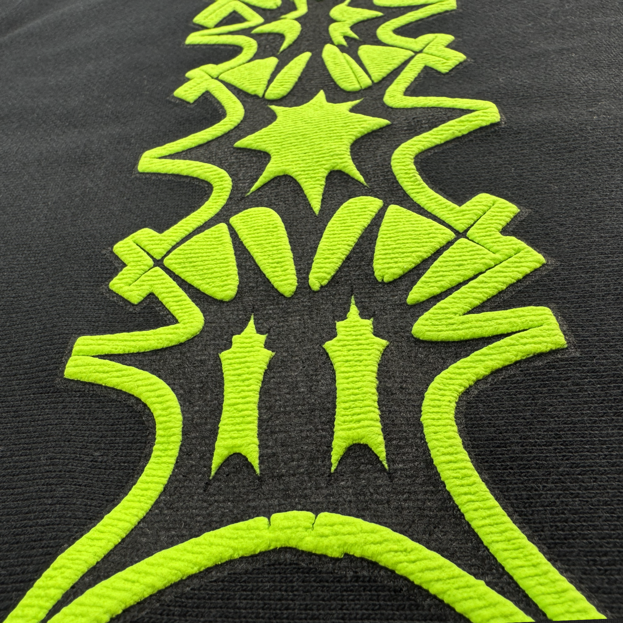Close-up of neon green embroidery on the black Distortion Hoodie by BOSK. Featuring symmetrical abstract shapes like stars and waves, the design creates vibrant contrast. The textured threads enhance depth and detail on the pigment-dyed cotton fleece, showcasing an intricate artistic motif.