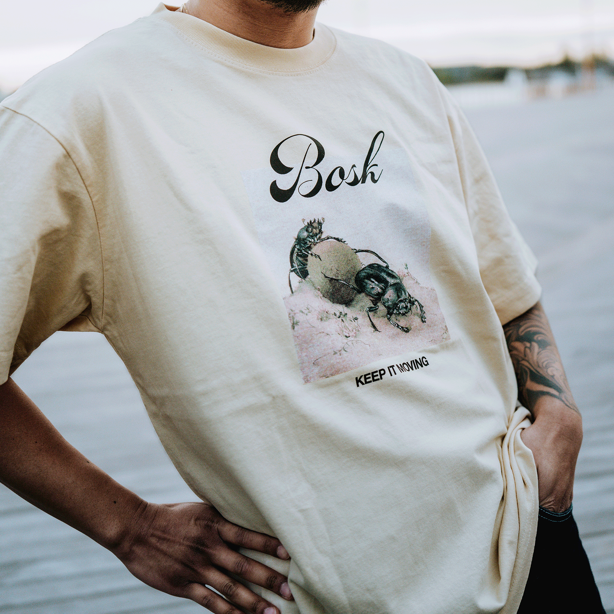 A person wears the BOSK Moving Tee, a classic fit light beige heavy-weight cotton shirt with two beetles on a rock, “Bosh” script above, and “KEEP THINKING” in all caps below. Hands rest on hips outdoors.