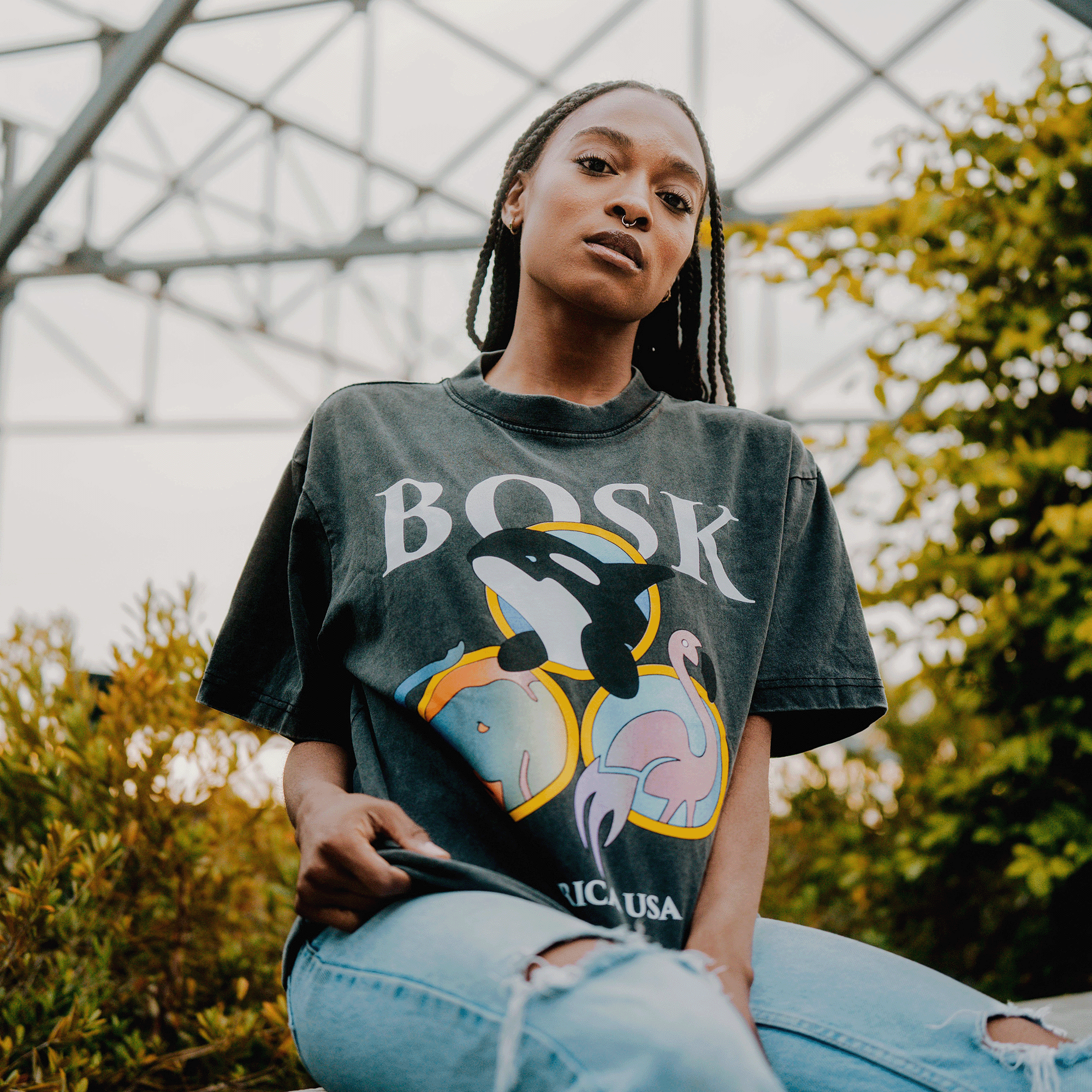 A young woman with long braids wears the BOSK World Tee, a classic fit, faded black heavyweight cotton shirt featuring an orca, flamingos, and "BOSK." She sits outdoors among greenery and metal, looking confidently at the camera.