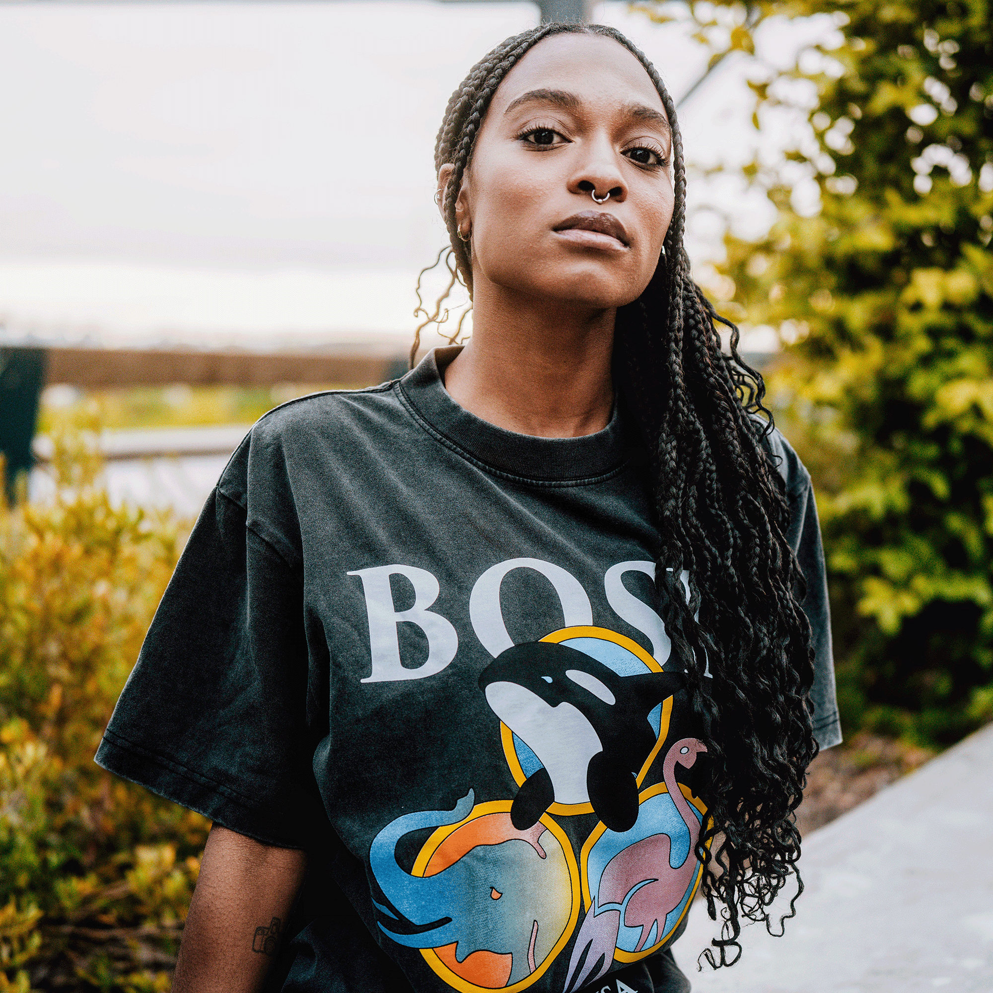 A person with long braided hair stands outdoors, wearing the BOSK Bosk World Tee, a faded black heavyweight cotton classic fit t-shirt featuring “BOSK” and colorful orca, elephant, and rhinoceros graphics. Green foliage and a blurred path are in the background.