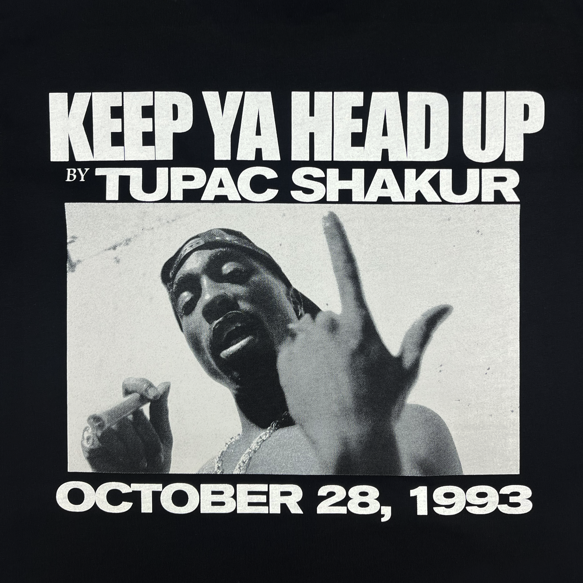 In a black and white image, a person holds a cigarette and gestures with one hand. Text above reads "KEEP YA HEAD UP BY TUPAC SHAKUR" and below "OCTOBER 28, 1993." This design is featured on the Brighter Days Tee by Oaklandish, celebrating the legacy of Tupac Shakur as part of their hip-hop collection.