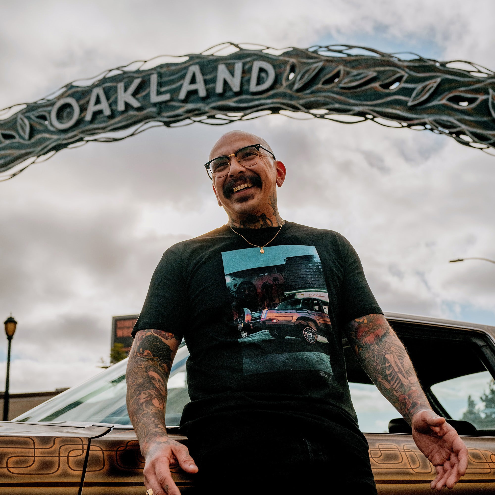 Man standing under an Oakland sign wearing a black t-shirt with an image of Armando’s lowrider car parked in front of a Nipsey Hussle mural.