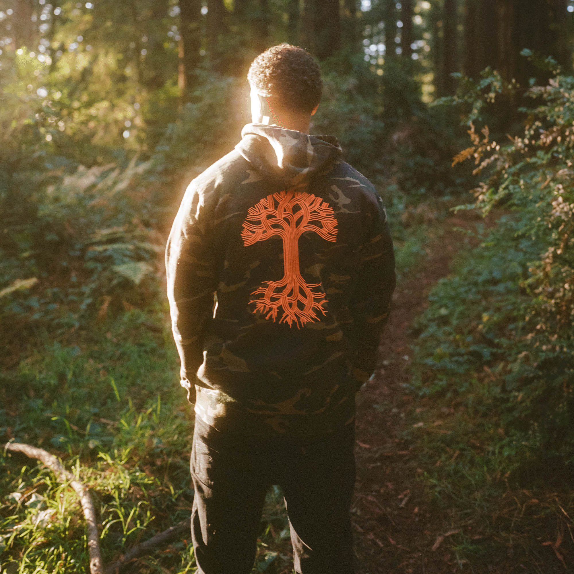 Back view of a model wearing camo pullover hoodie sweatshirt with a large orange Oaklandish tree logo in the center.