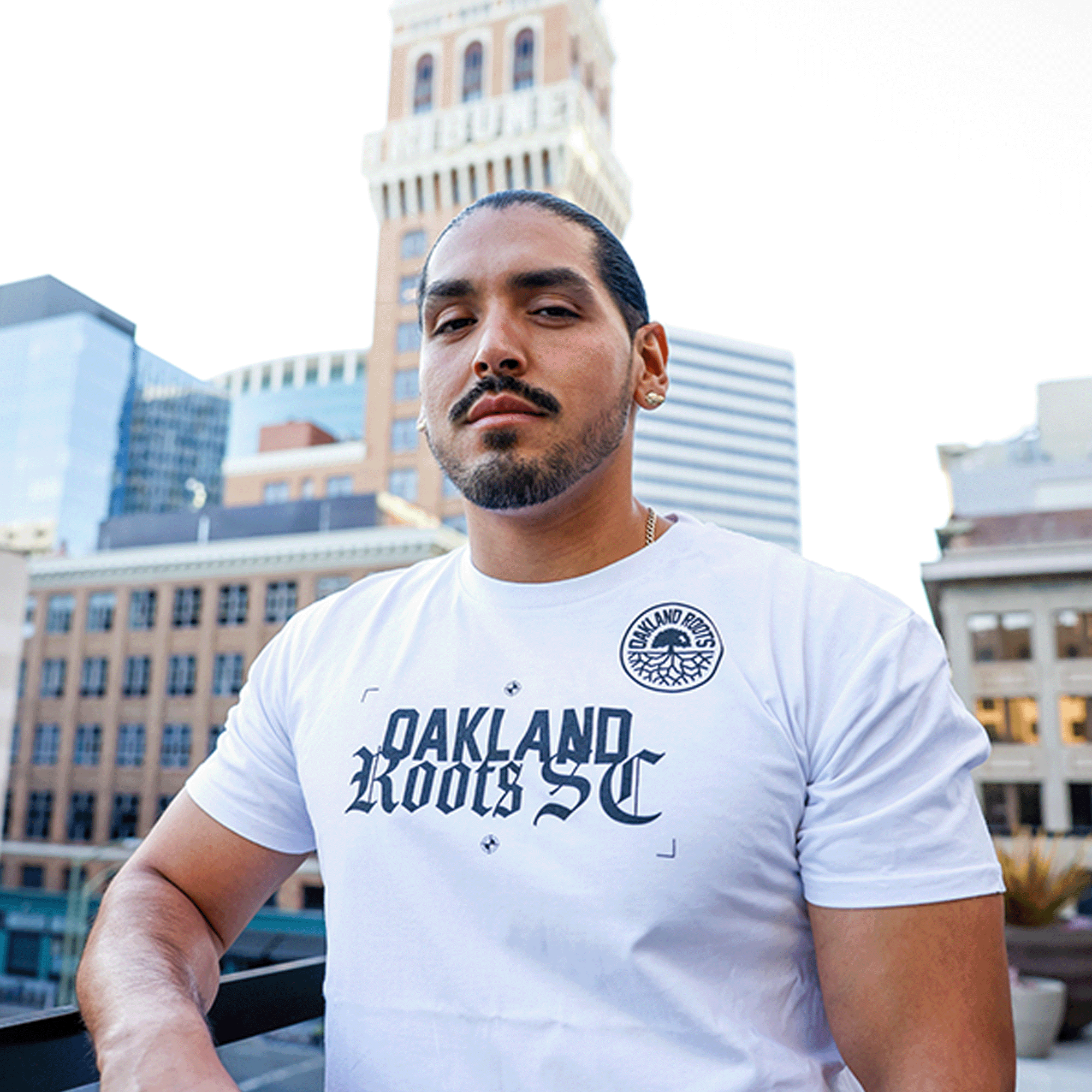 A man with long dark hair and a beard stands on a rooftop in an urban Oakland setting. He is wearing an Oakland Roots SC Futbol Town Tee and looking confidently at the camera. Behind him, there are several buildings, including a tall one with the word "TRIBUNE" partially visible at the top.