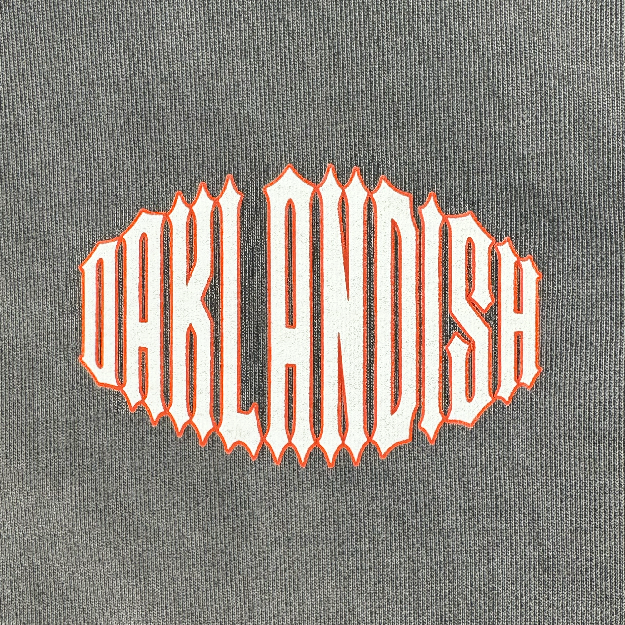The Chopper Heavy Hoodie by Oaklandish is a gray, oversized drop shoulder pullover made from 100% cotton fleece. It features "OAKLANDISH" in white text with bolded white and orange outlines, using a stylized font for a dynamic, edgy look centered on the garment.