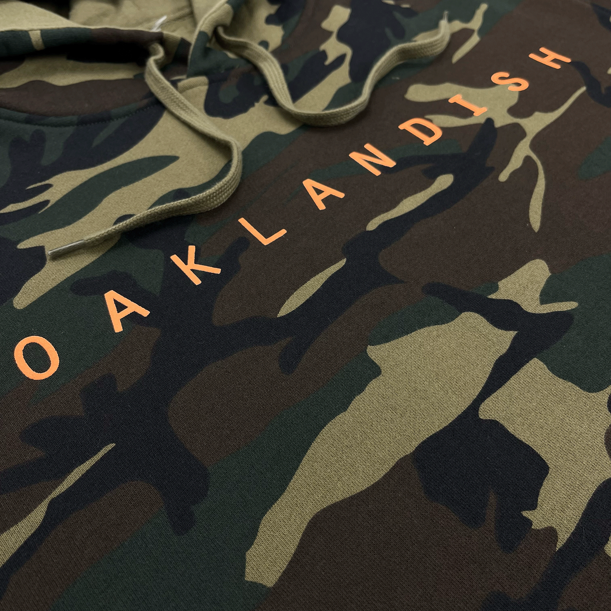 Large orange OAKLANDISH wordmark on the front chest on a camo pullover hoodie sweatshirt.