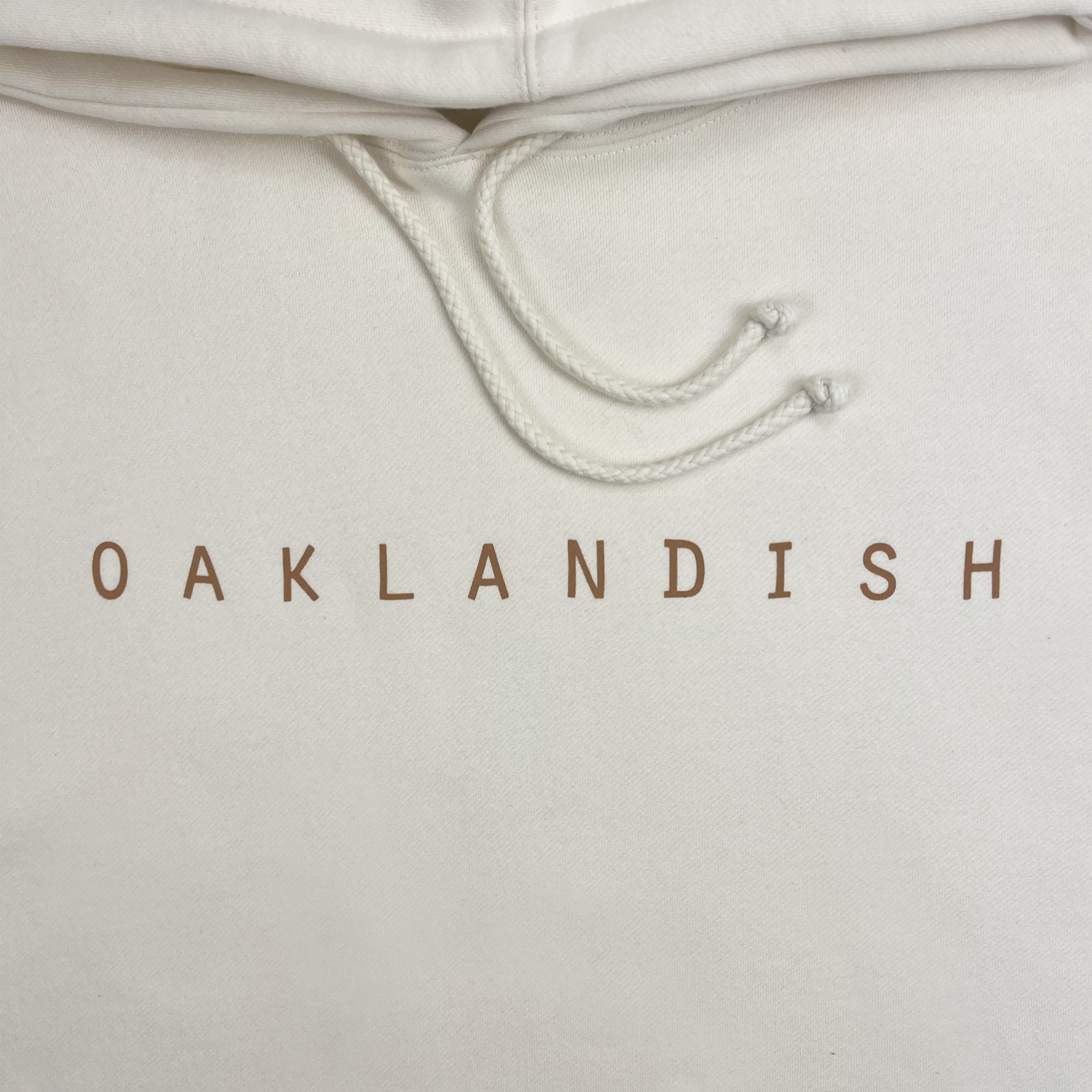 Close-up of the Oaklandish Classic Hoodie in white, featuring "OAKLANDISH" in bold brown, all-capital letters across the chest. This men's fit hoodie by Oaklandish has adjustable drawstrings tied in small knots at their ends, hanging from the hood. The fabric appears soft and well-finished.