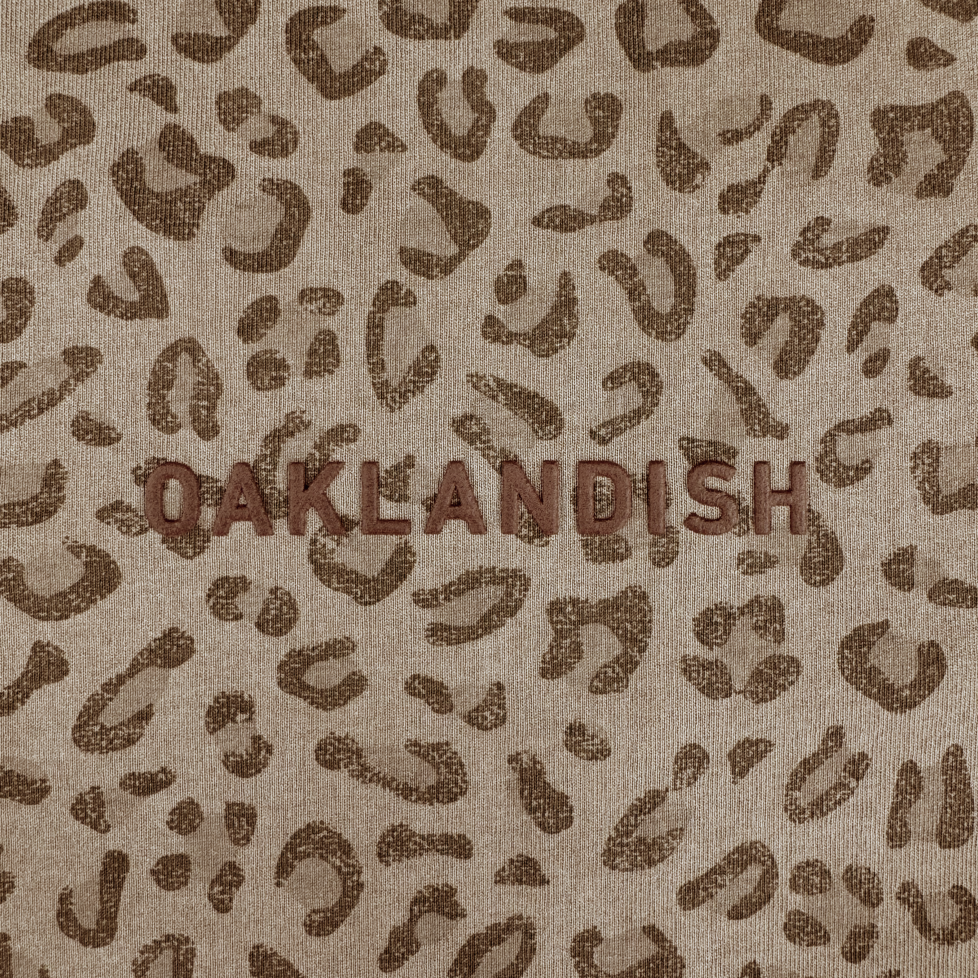 The Oaklandish Classic Logo Tee by Oaklandish features a tan base with brown spots, resembling a men's leopard print, and bold "OAKLANDISH" embroidery on the front. Crafted from heavyweight cotton for a textured look.