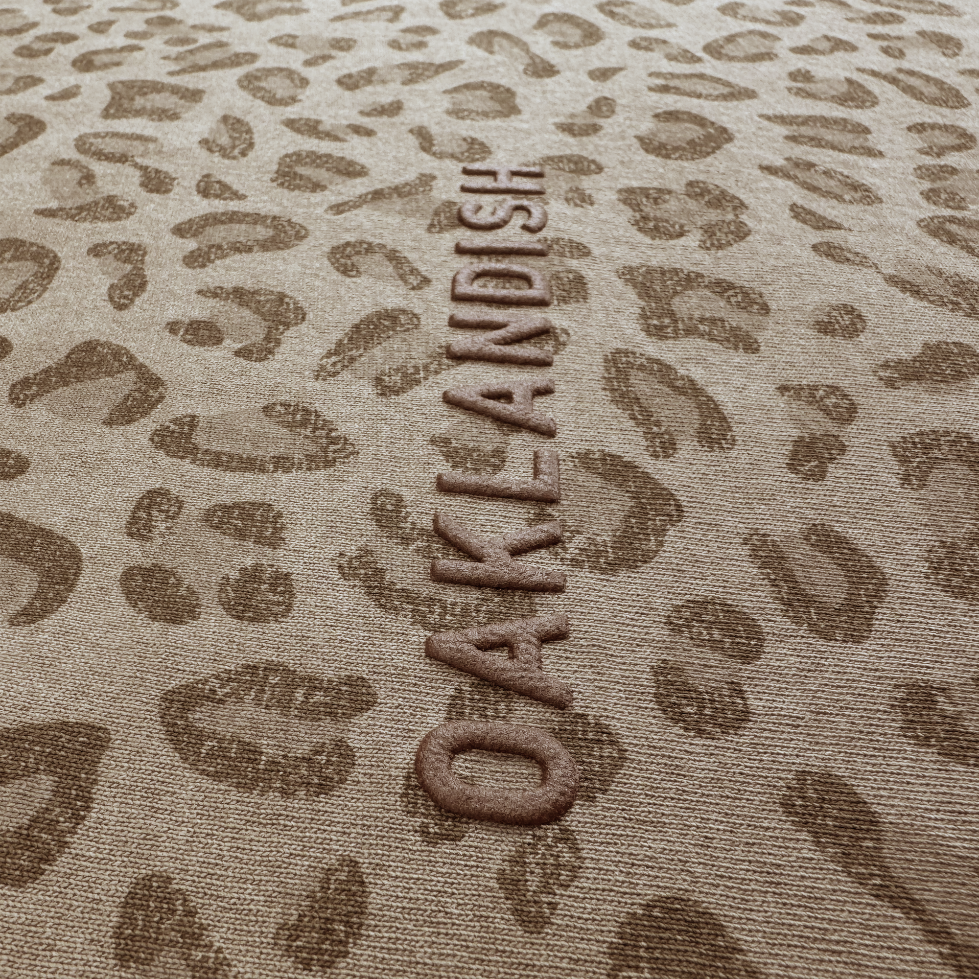 A close-up of heavyweight tan cotton with a brown leopard print and "OAKLANDISH" embossed vertically, perfectly matching the Oaklandish Classic Logo Tee’s color scheme.