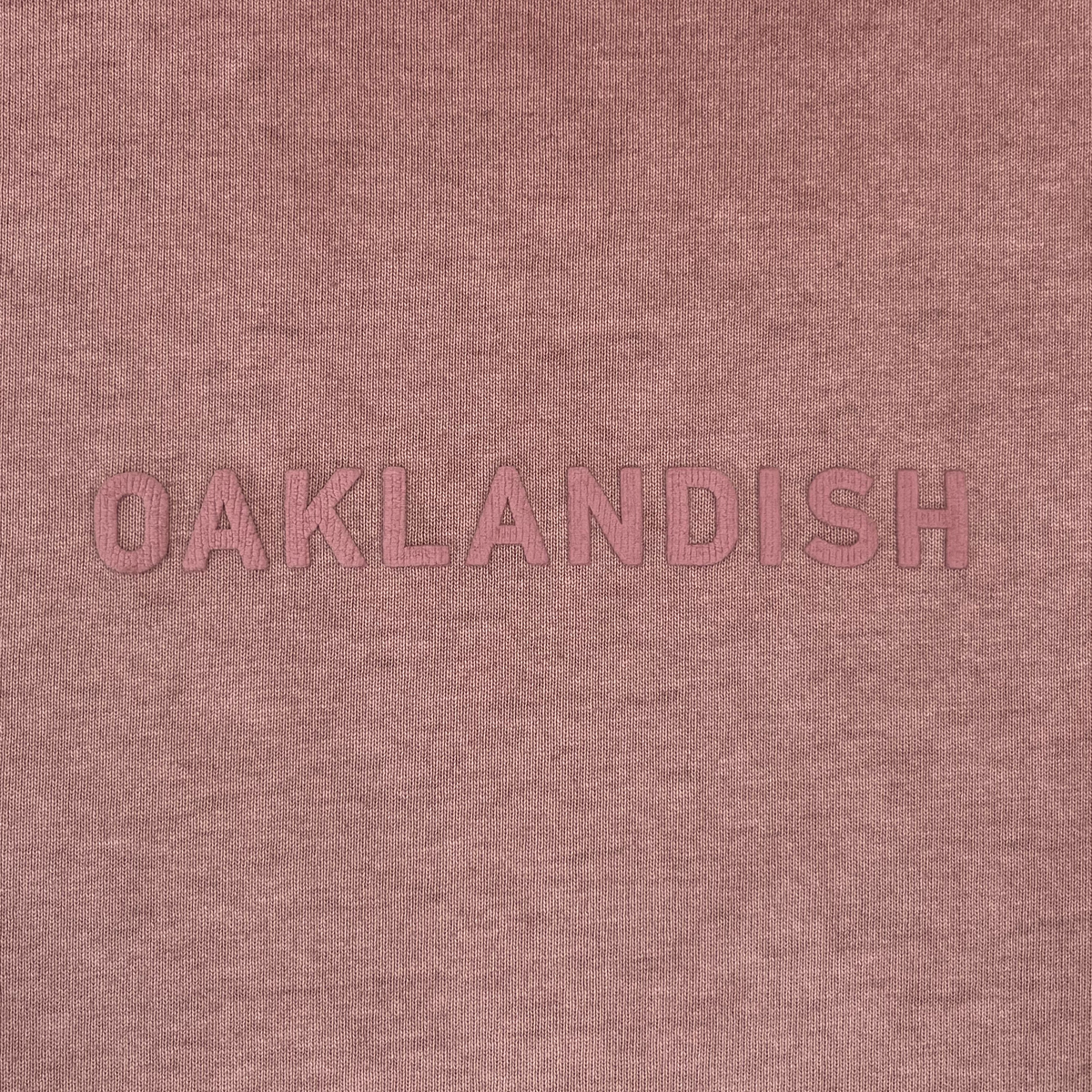The Oaklandish Classic Logo Tee by Oaklandish features bold, uppercase “OAKLANDISH” lettering in pink, centered on a soft, textured dusty rose or mauve heavyweight cotton background. The minimalist, monochromatic design offers subtle contrast and comes in a classic fit men’s sizing.