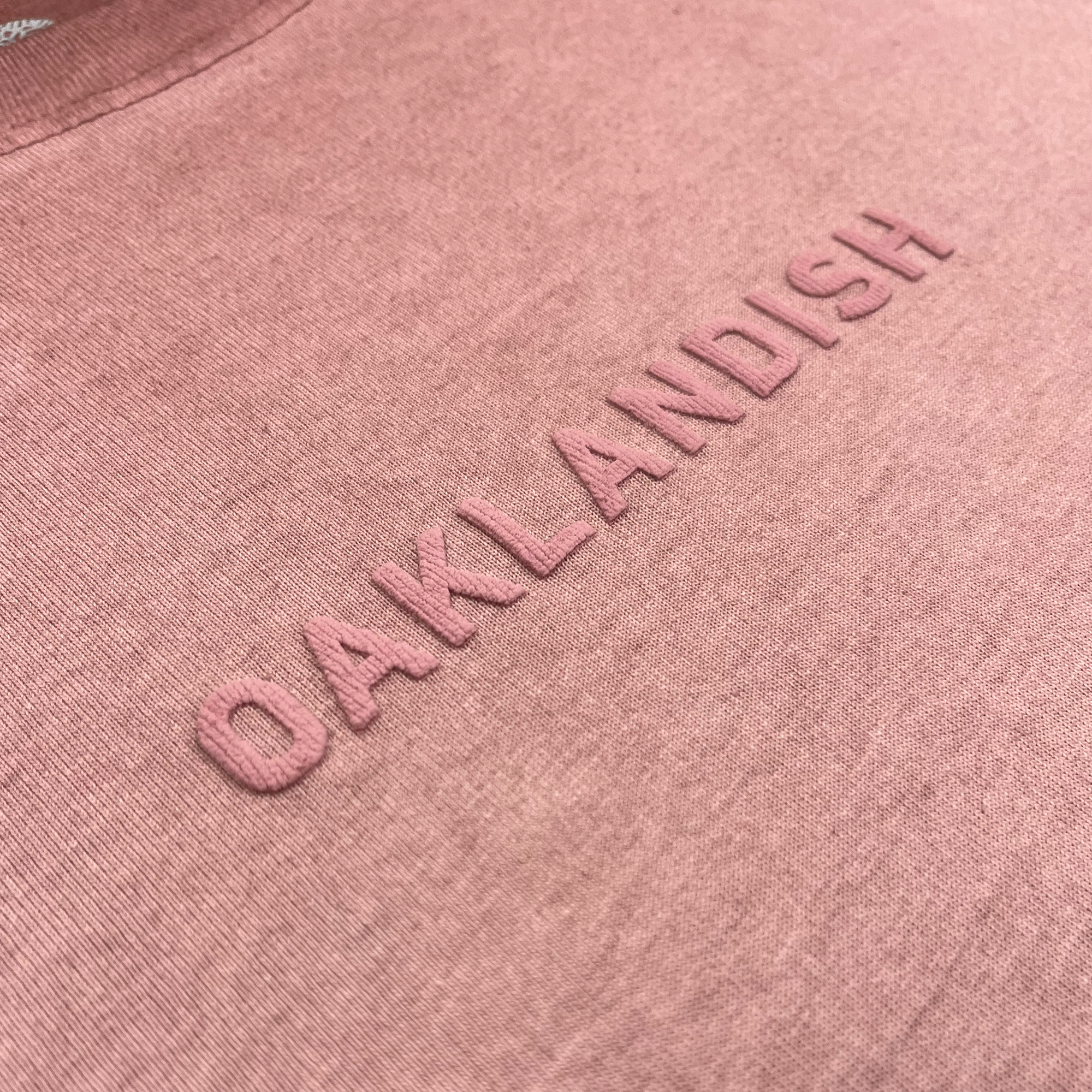 A close-up of the Oaklandish Classic Logo Tee by Oaklandish, showcasing heavyweight pink cotton fabric with “OAKLANDISH” embroidered in raised, matching pink letters across the chest. The detailed texture and stitching emphasize its quality and classic men’s sizing.