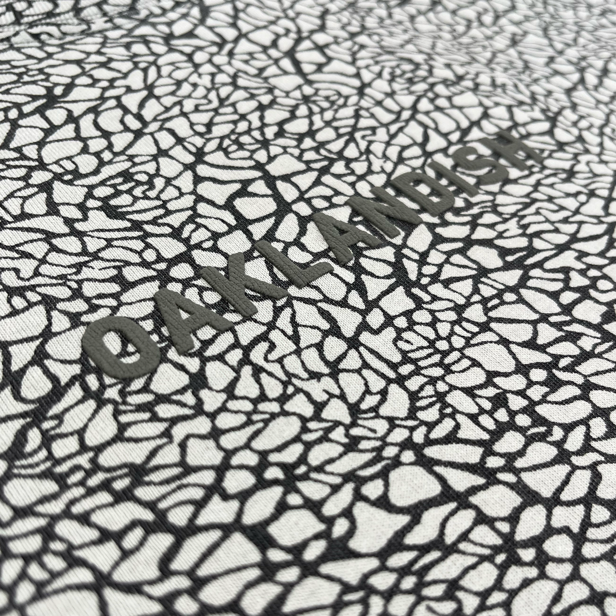 Close-up of the Oaklandish Classic Logo Tee’s textured surface, featuring an irregular, black-and-white cracked pattern. “OAKLANDISH” is prominently embossed across the center in bold, uppercase letters with a distinctive puff print technique that partially merges with the intricate background—showcasing the signature style and heavyweight cotton of this classic fit tee from Oaklandish.