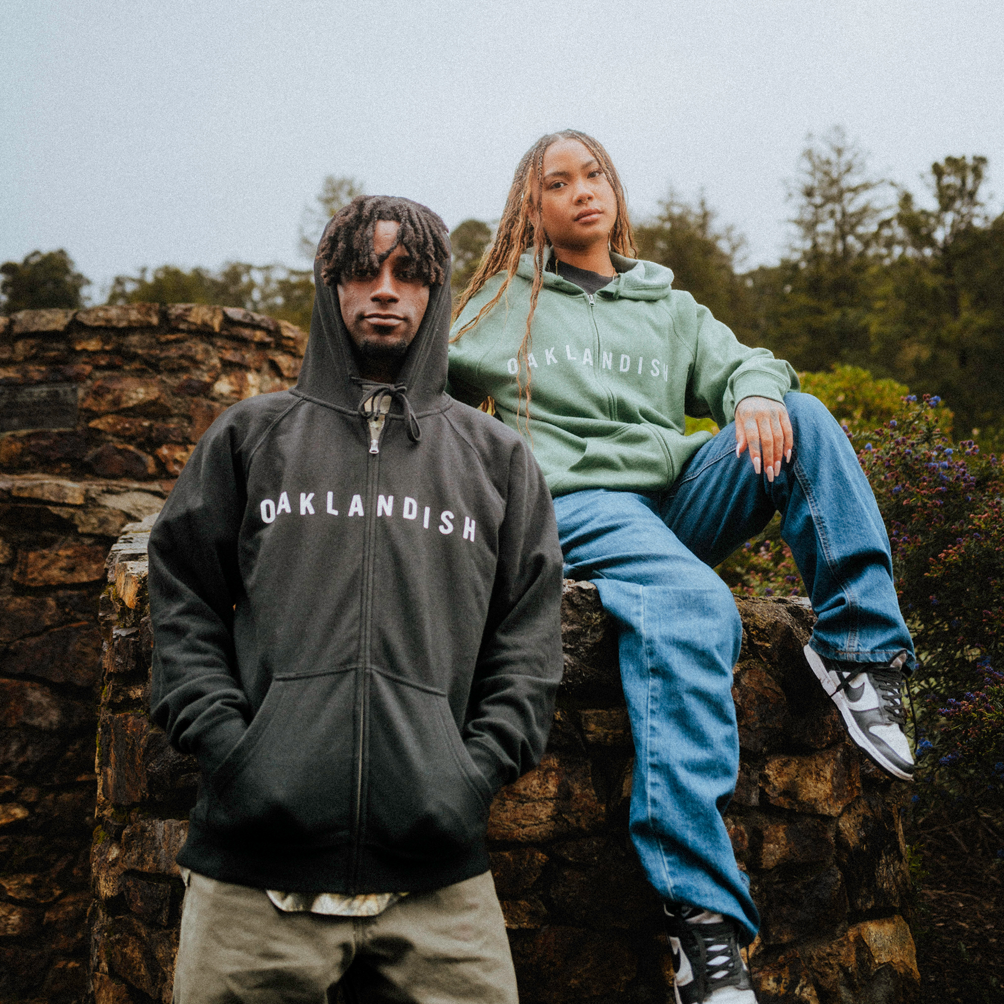 Two individuals in "Oaklandish Classic Zip Hoodie" apparel pose on a stone structure surrounded by nature. One stands in a black hoodie, the other sits casually in green. Both exude confidence facing the camera, complemented by trees and shrubs as their perfect unisex backdrop.