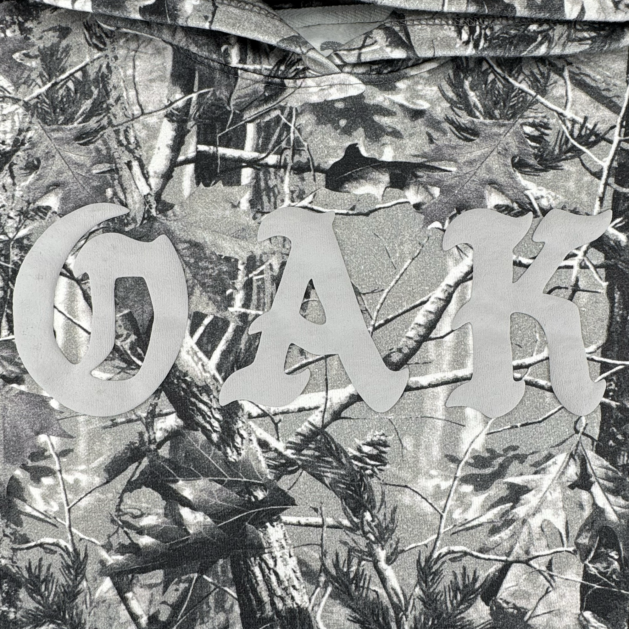 The Oaklandish Cork Hoodie features a grey wood camo design with forest-inspired branches, leaves, and pine cones. Bold "OAK" lettering in light gray makes this men’s outerwear stylish and unique.