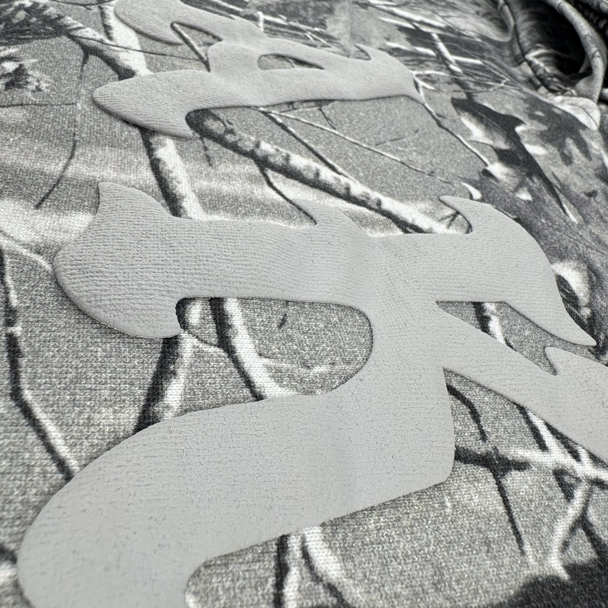 Close-up of the Oaklandish Cork Hoodie fabric, showing grey wood camo with raised matte patches forming abstract or Asian-inspired characters. Textured like tree branches, this adds unique dimension to the men's outerwear.