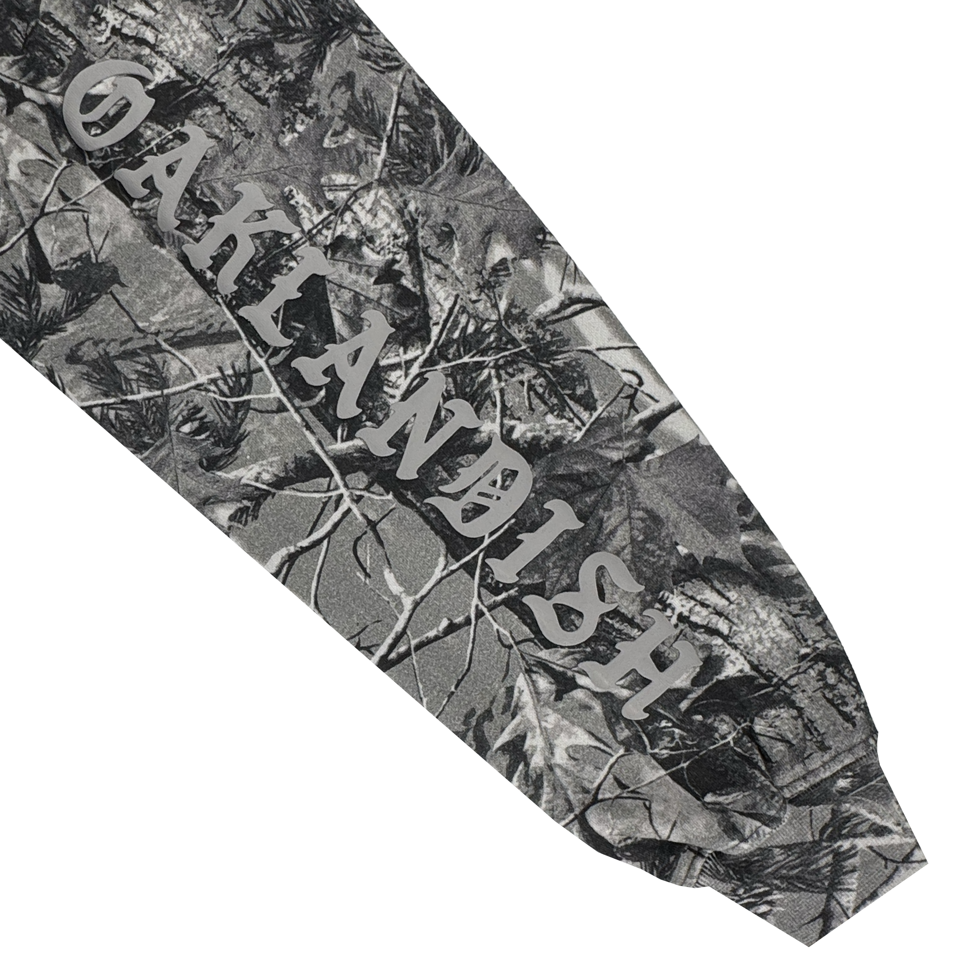 A close-up of the Cork Hoodie by Oaklandish shows a grey wood camo forest pattern. "OAKLANDISH" is printed in large, white serif letters diagonally across the men's outerwear sleeve, surrounded by tree branches and leaves.