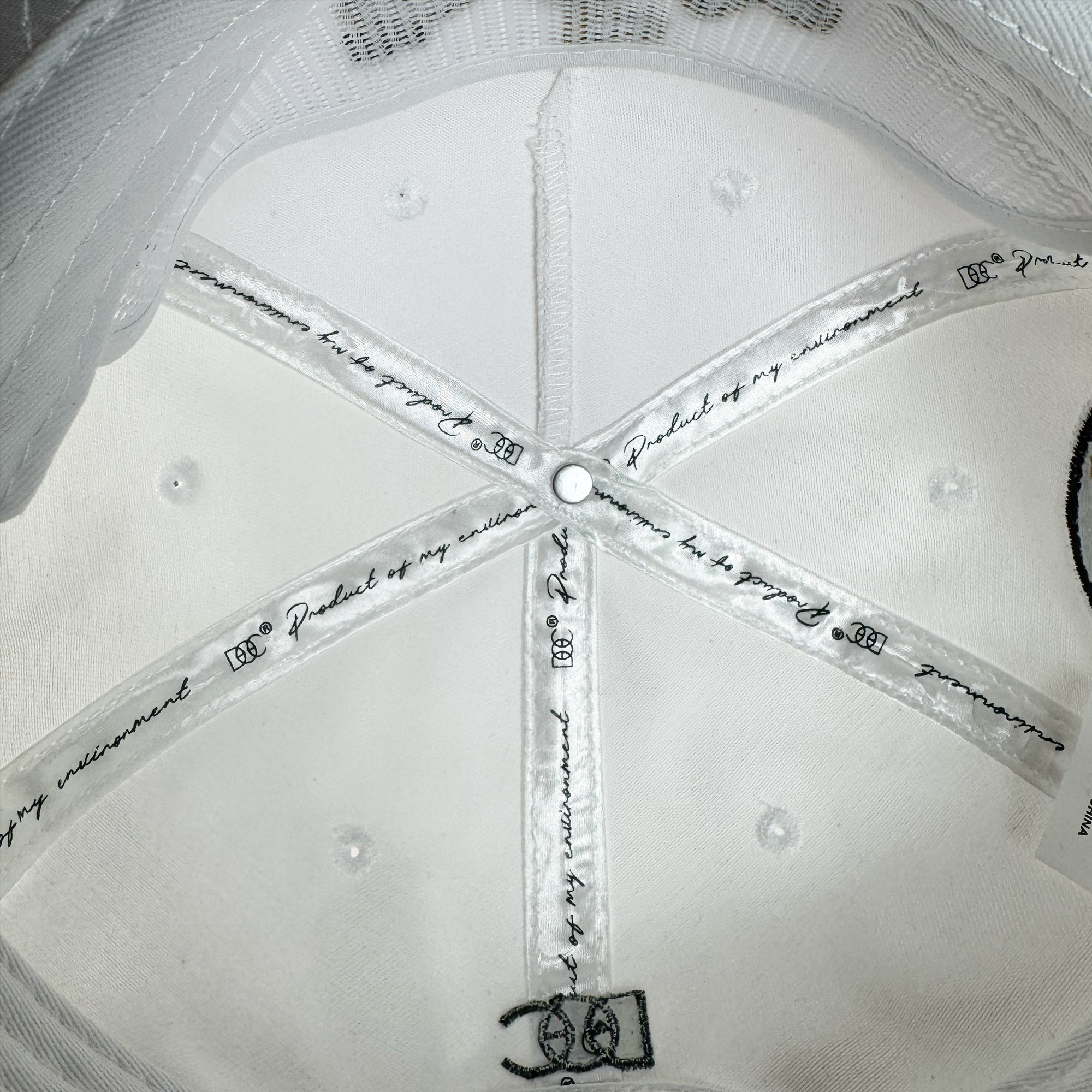 Close-up of the inside top of the white Bay Area 5 Panel By DOC hat by Oaklandish, showing intersecting seams with printed text and "DC" above the band. A small rivet at the seam intersection highlights stitching details.