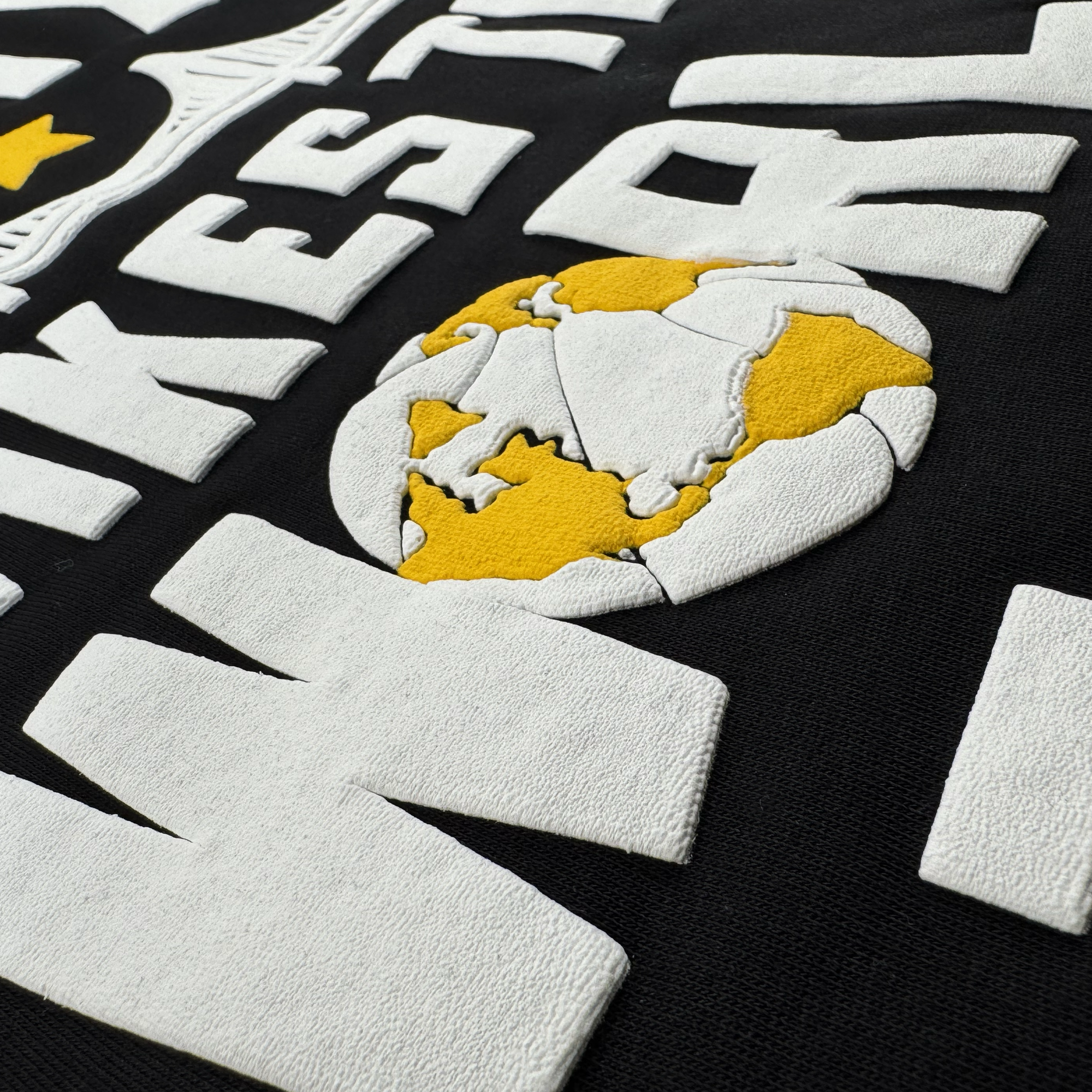 Close-up of raised, textured white letters and a yellow-and-white globe graphic on black fabric. The design, from the Oaklandish Bay Bridge Hoodie By DOC V2, shows simplified continents and a partially visible yellow star above.