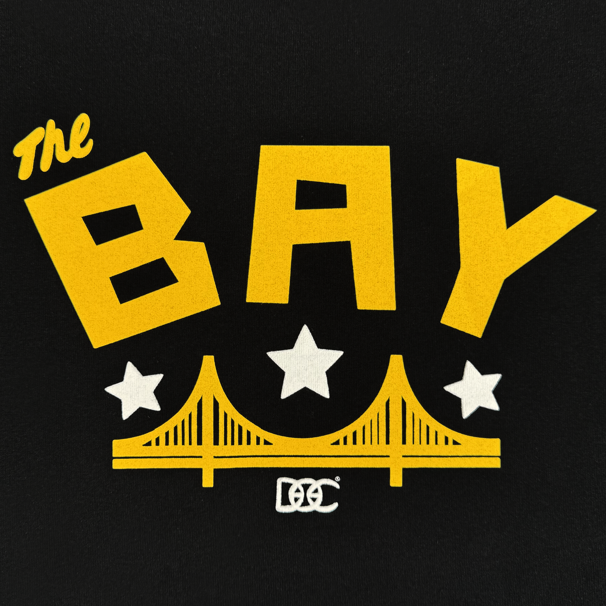 Bold yellow text reads "The BAY" above a stylized yellow bridge and three white stars on black. Below, white interlocking “DC” letters complete the look of the Bay Bridge Hoodie By DOC V2 from Oaklandish—perfect for any hoodie.