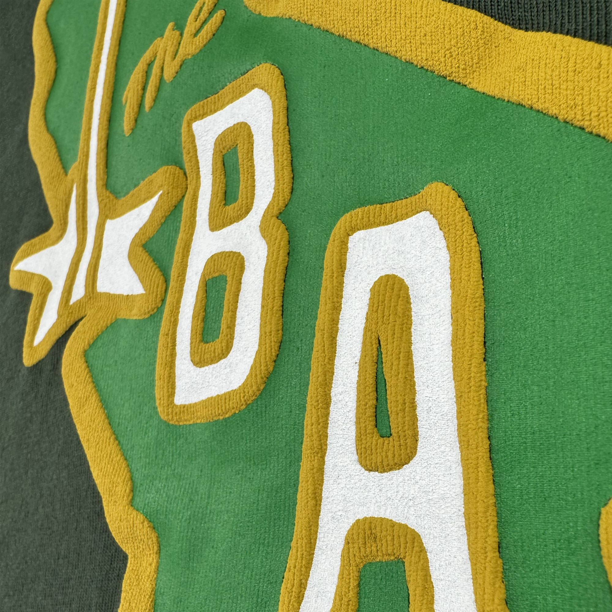 A close-up of textured embroidery on the CA Bay Area Tee By DOC by Oaklandish shows “THE BAY” in white and gold letters on green fabric, with a gold landmass outline, white star, and raised stitching for a 3D effect.
