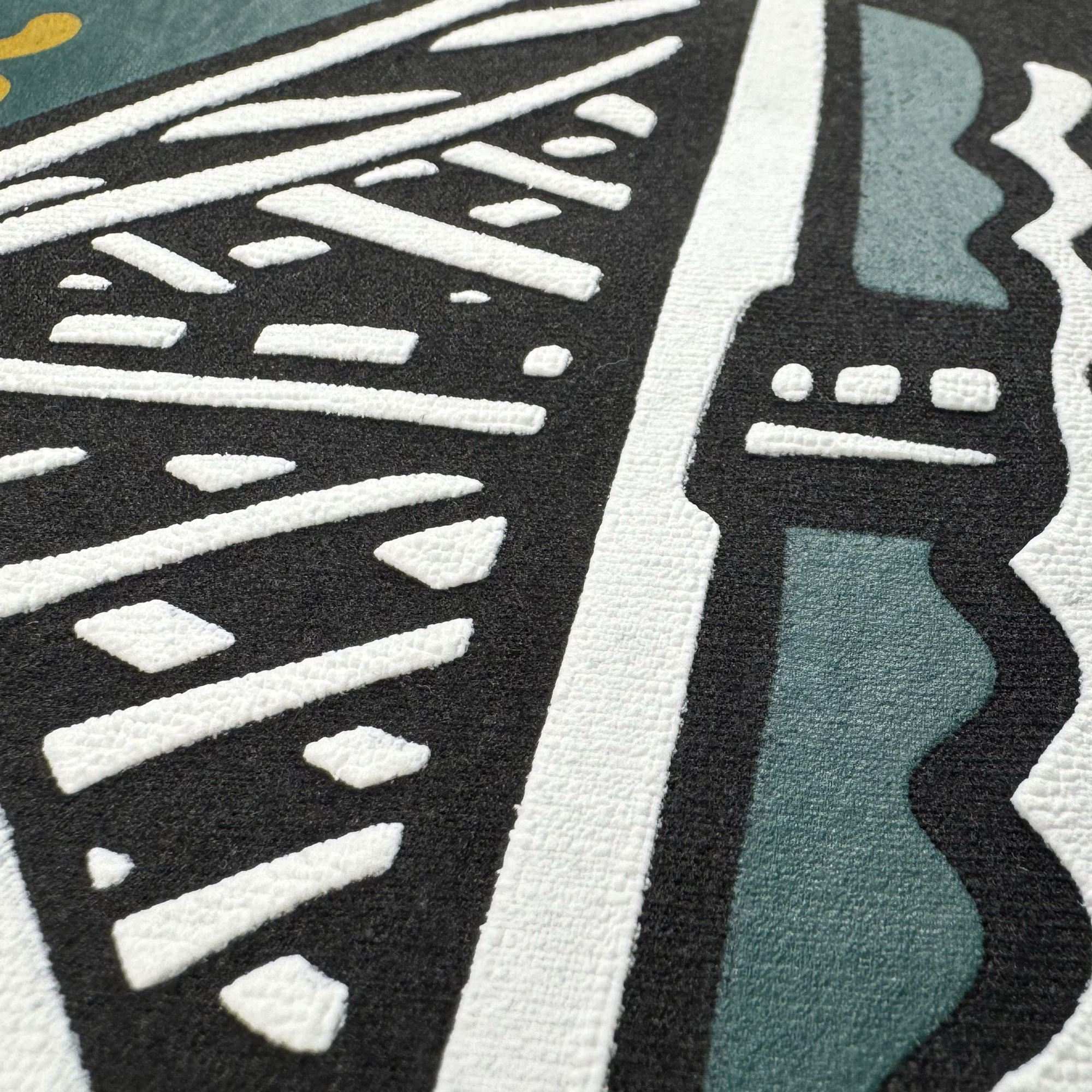 A close-up of the Bay Area CC Tee by Oaklandish shows bold black, white, and teal shapes with angular lines, crosshatch patterns, and yellow dots, creating an abstract design inspired by a stylized bridge or architecture.