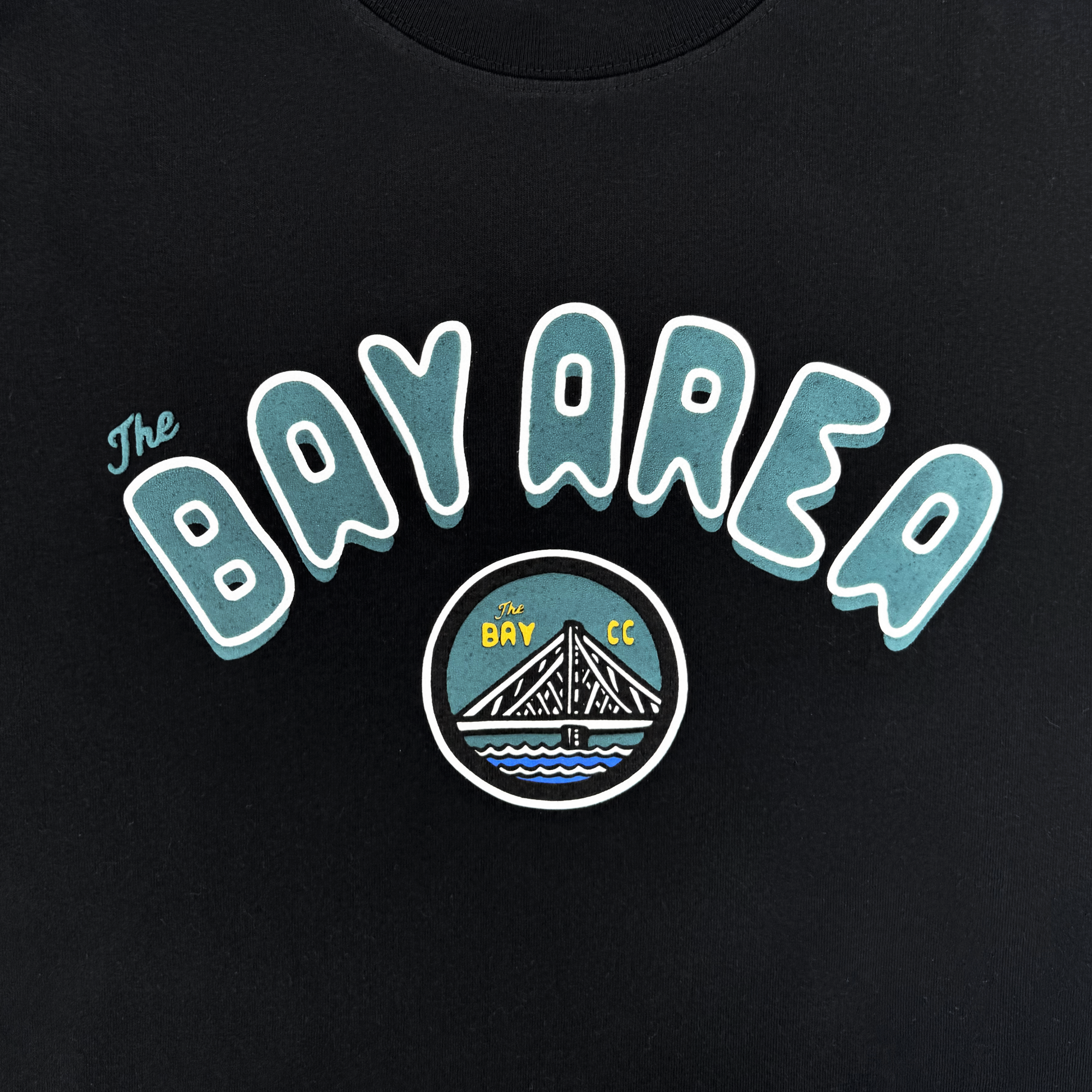 The Oaklandish Bay Area CC Tee By DOC is a black heavyweight cotton classic fit shirt with "The Bay Area" in bold blue bubble letters above a round patch showing a bridge, water, sun, and "The Bay" and "CC" in yellow. Part of The Bay Collection.