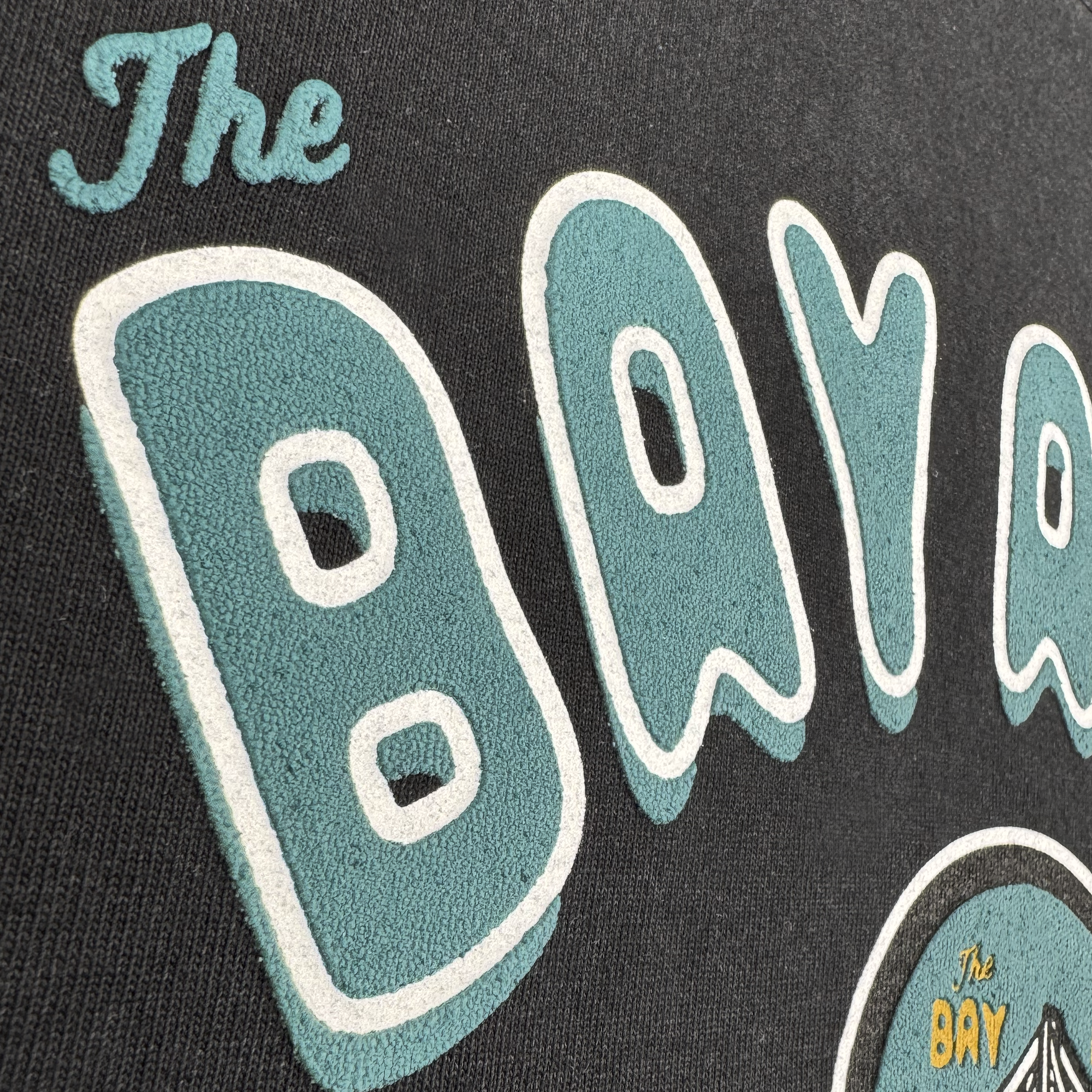 Close-up of textured "Bay Area CC" lettering in retro teal and white on black fabric. The Oaklandish Bay Area CC Tee by DOC is a classic-fit, heavyweight cotton t-shirt with a partially visible circular logo. Part of The Bay Collection.