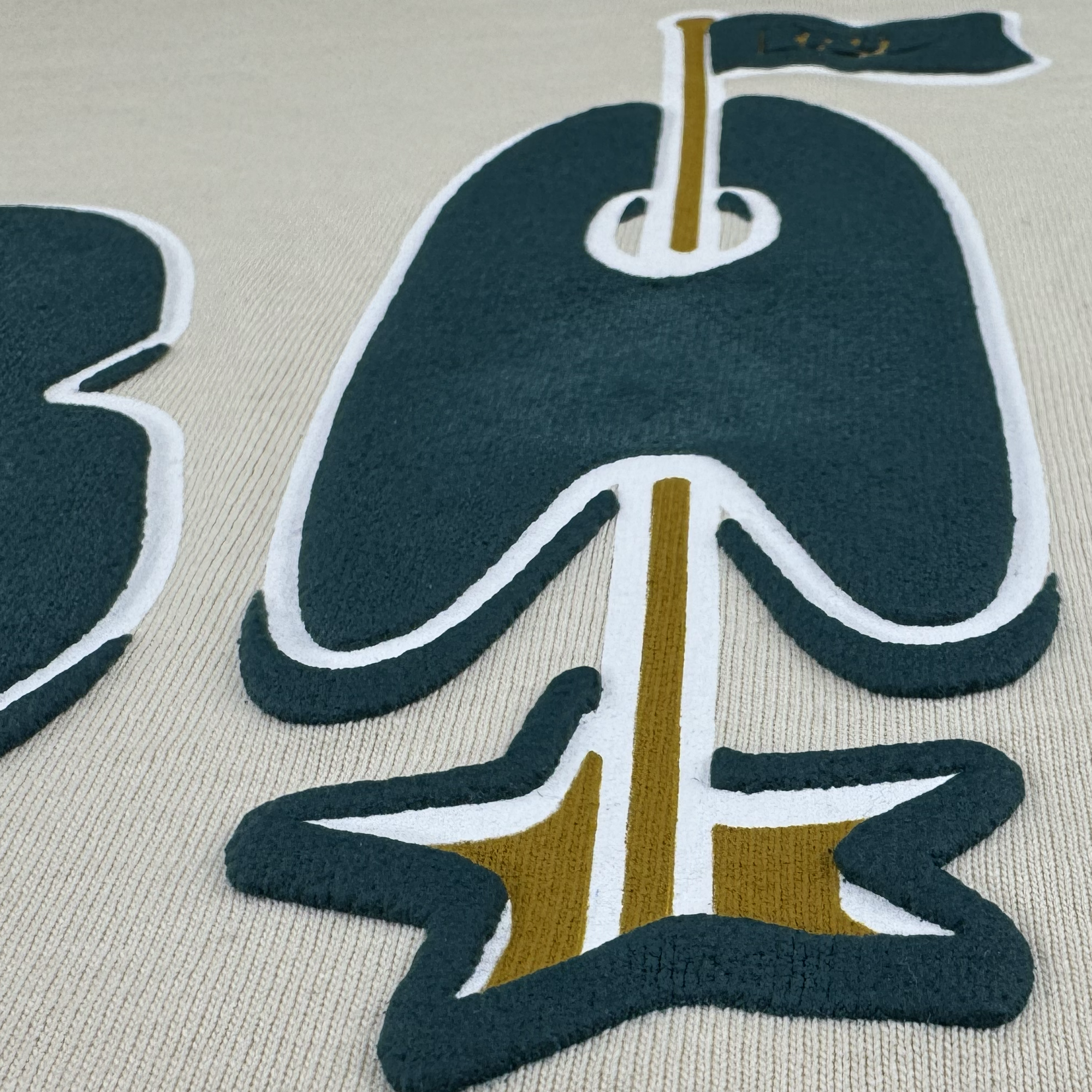 A close-up of textured embroidery on heavyweight cotton shows a stylized dark green "A" with a gold and white arrow ending in a star, set on a light beige knit classic fit The Bay Flag Tee By DOC by Oaklandish.