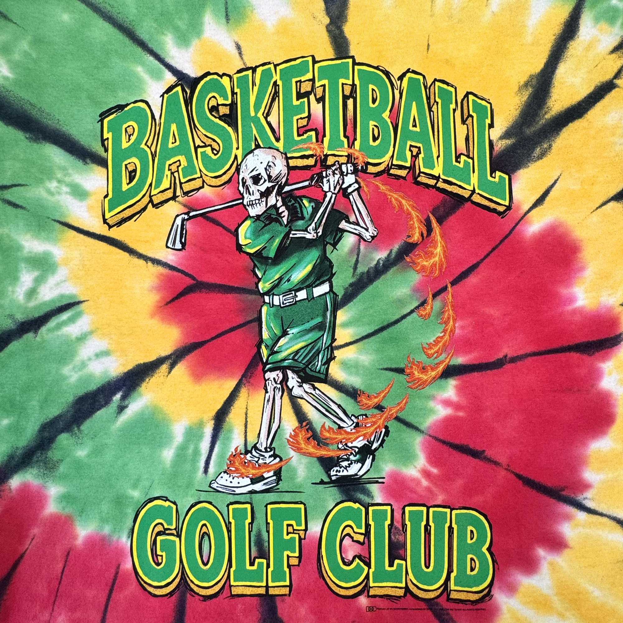 A green skeleton swings a flaming golf club while wearing fiery golf shoes, set against a yellow, red, and green tie-dye background. “BASKETBALL” is printed above and “GOLF CLUB” below. This heavyweight cotton classic fit t-shirt is called The Dead Golf Tee By DOC from By DOC.