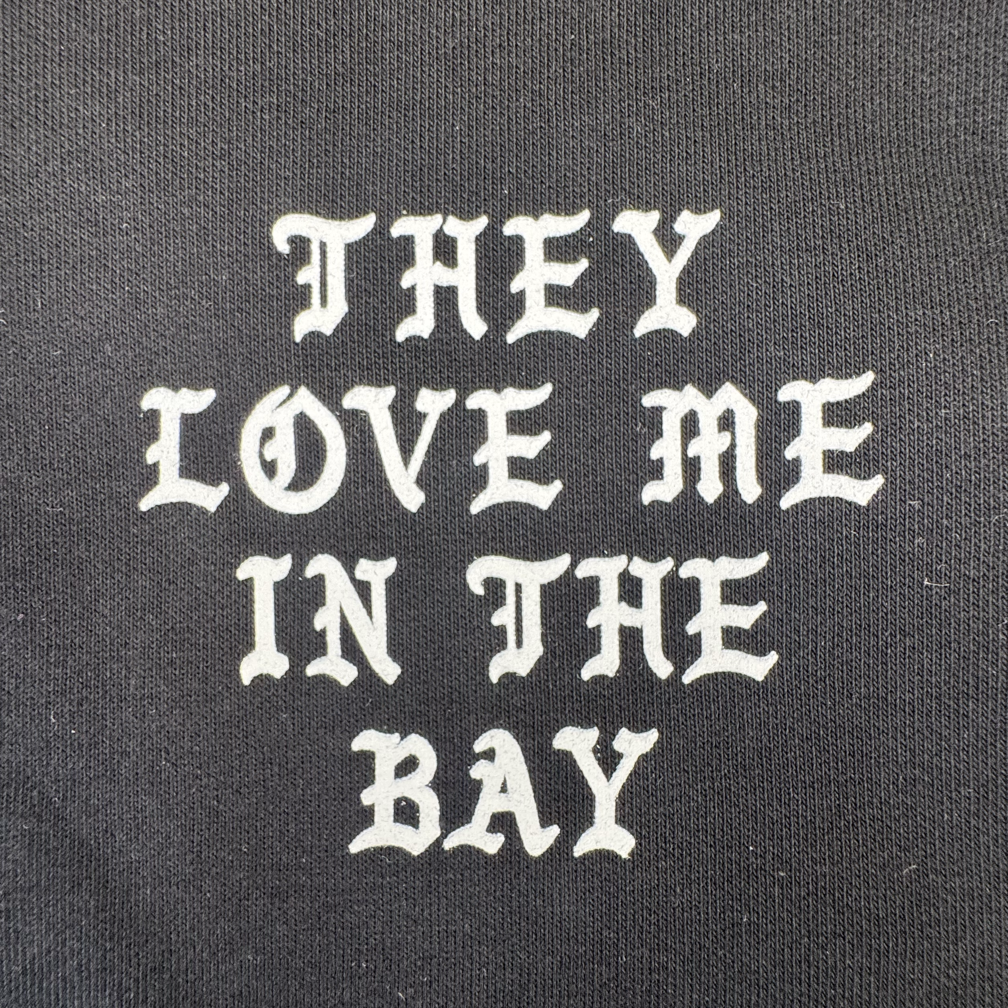 Sure! Here’s a rewritten version using your product data: Text on black fabric reads: “THEY LOVE ME IN THE BAY” in bold, white, Old English-style font. The textured background suggests a heavyweight fleece construction from the By DOC They Love Me Zip—perfect for a cozy shirt or zip-up hoodie.