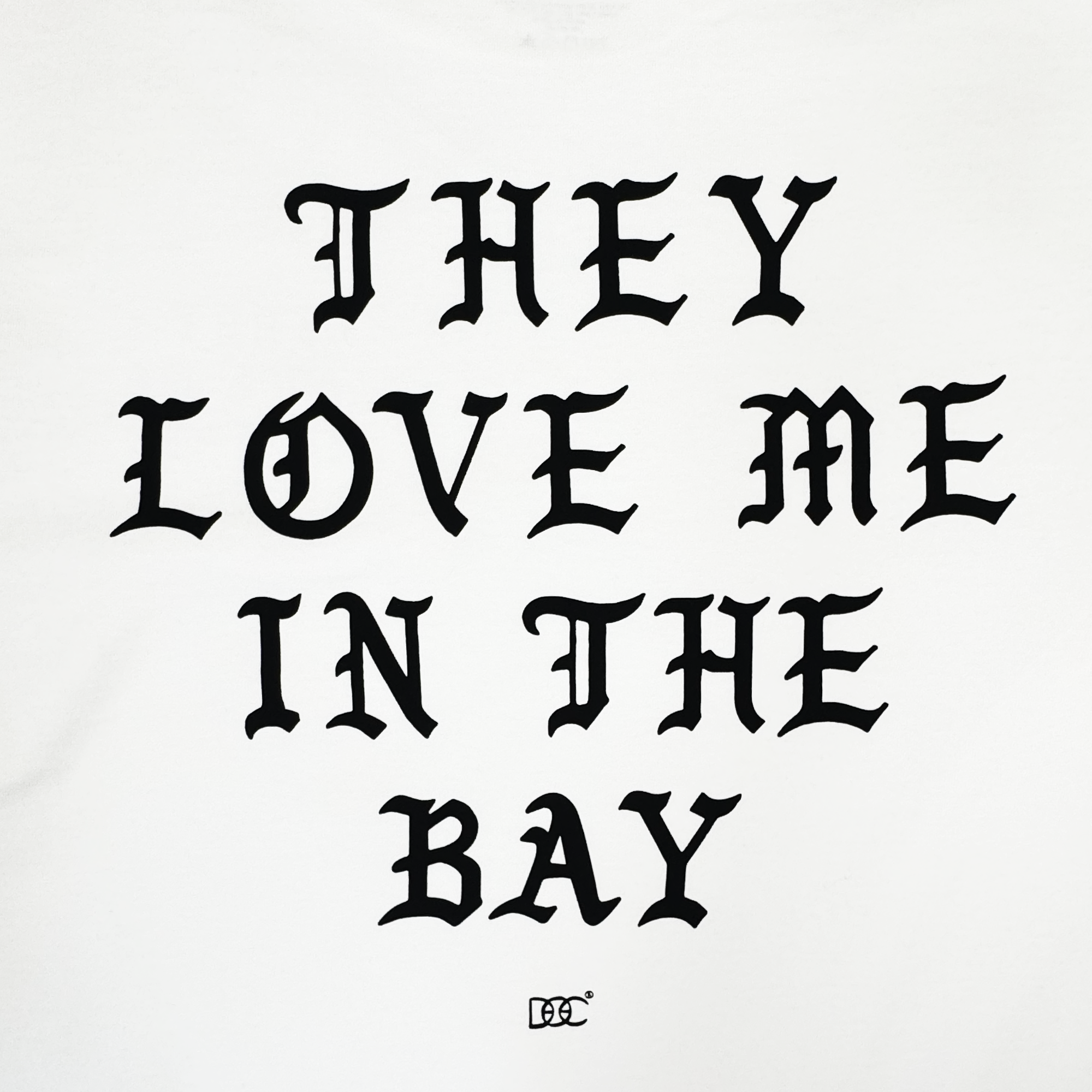 The They Love Me LS Tee By Doc from By DOC features bold, black Gothic-style text in all uppercase letters on a crisp white background, reading “THEY LOVE ME IN THE BAY.” A small By DOC logo with two interlocking C’s is centered beneath the phrase. This simple yet striking design is printed on a heavyweight cotton long sleeve t-shirt for durability and lasting comfort, and is available in men’s sizing.