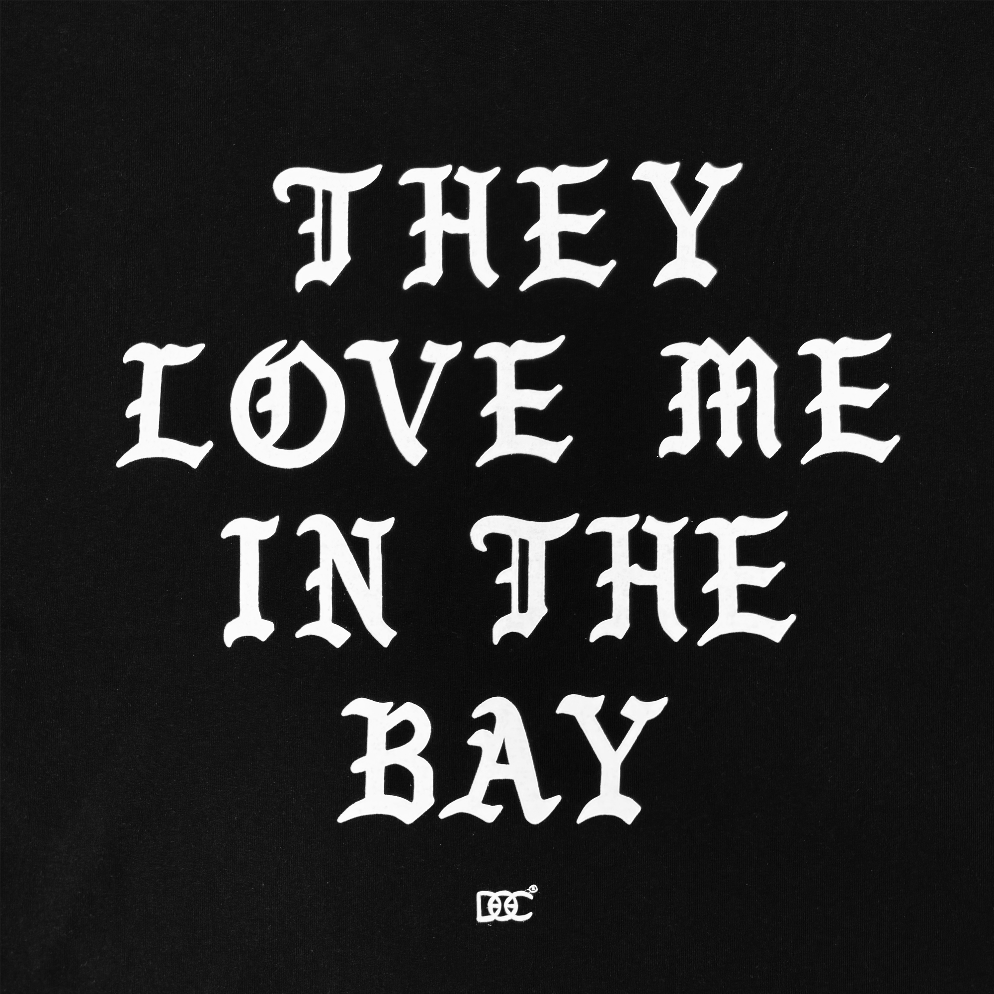 White text on a black background reads “THEY LOVE ME IN THE BAY” in bold, gothic-style uppercase letters. The design appears on the They Love Me Tee By Doc from By DOC’s Bay Collection—a classic fit t-shirt made from heavyweight cotton. A small decorative symbol is centered near the bottom of the artwork.