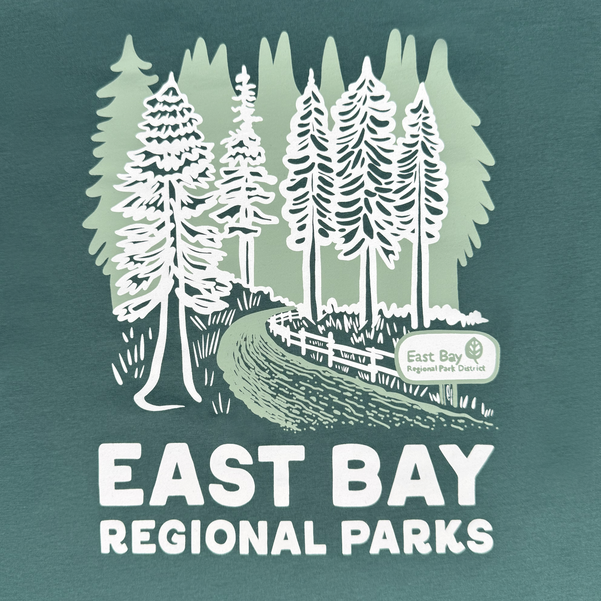 Featuring a woodland scene with stylized trees on green, a path, a wooden fence, and an "East Bay Regional Park District" sign, this Women's East Bay Regional Parks Tee by Oaklandish boasts bold white "EAST BAY REGIONAL PARKS" text. Made from 100% cotton.