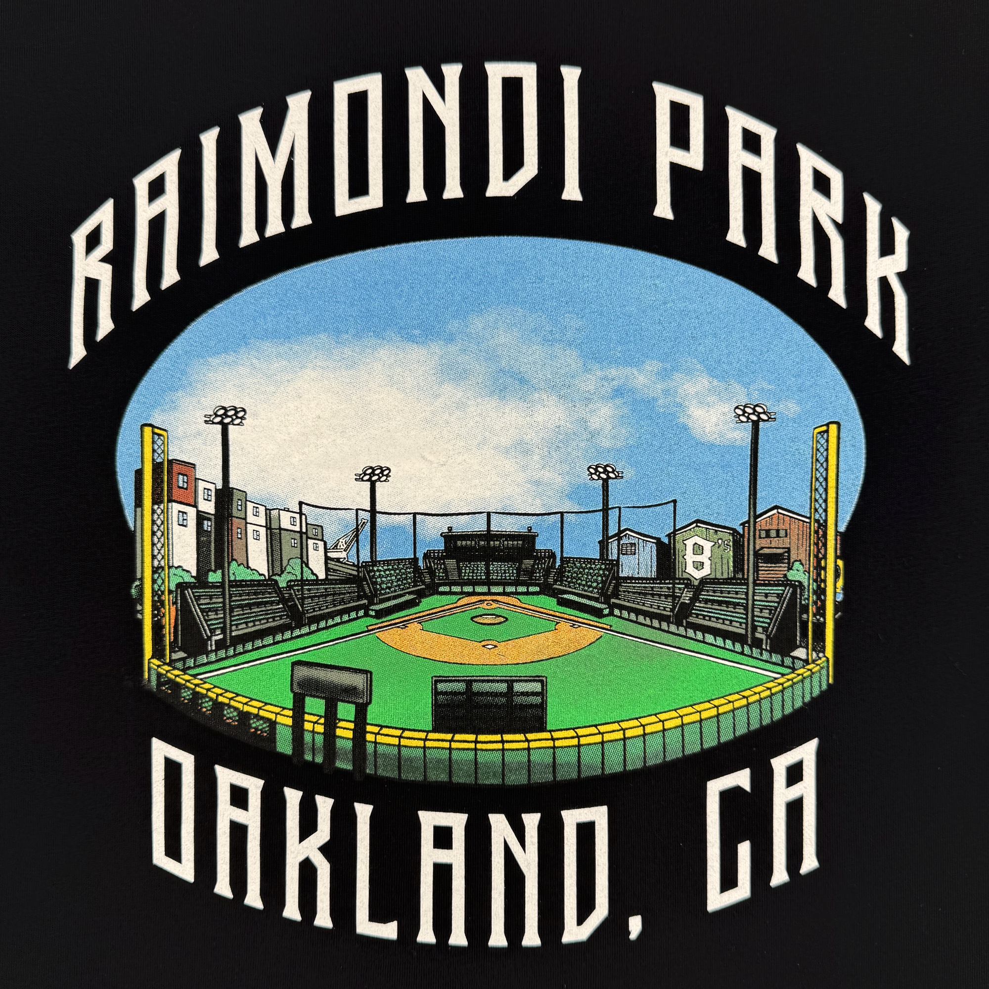 The Fisheye Tee by Oaklandish Ballers features an illustration of Raimondi Park in Oakland, CA—home of the Oakland Ballers—with green bleachers, city buildings, and light poles under a blue sky.