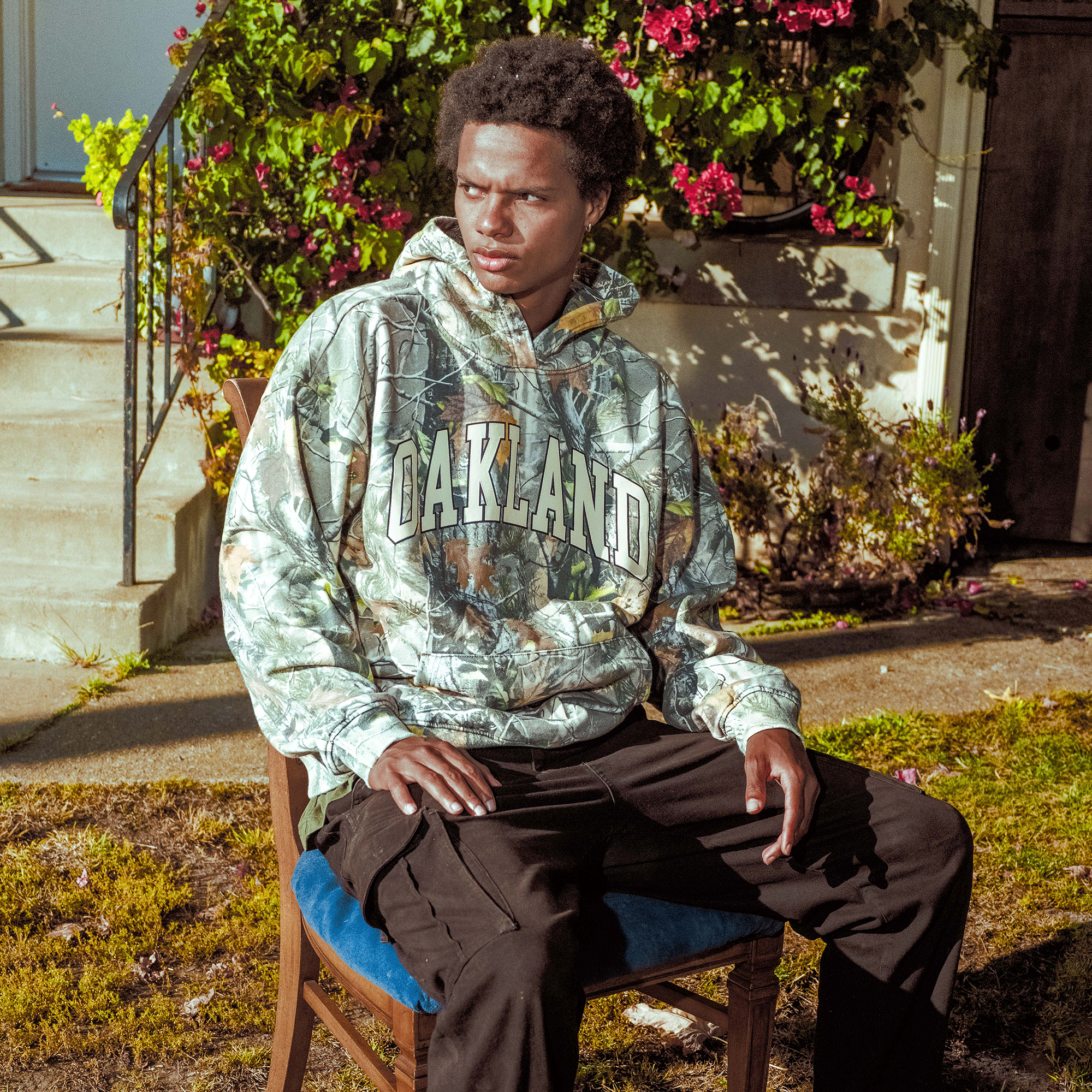 A young man sits outside on a wooden chair, wearing the Oaklandish Foliage Hoodie and black pants. Sunlight highlights his face as greenery and flowering bushes frame the concrete steps by the building entrance.