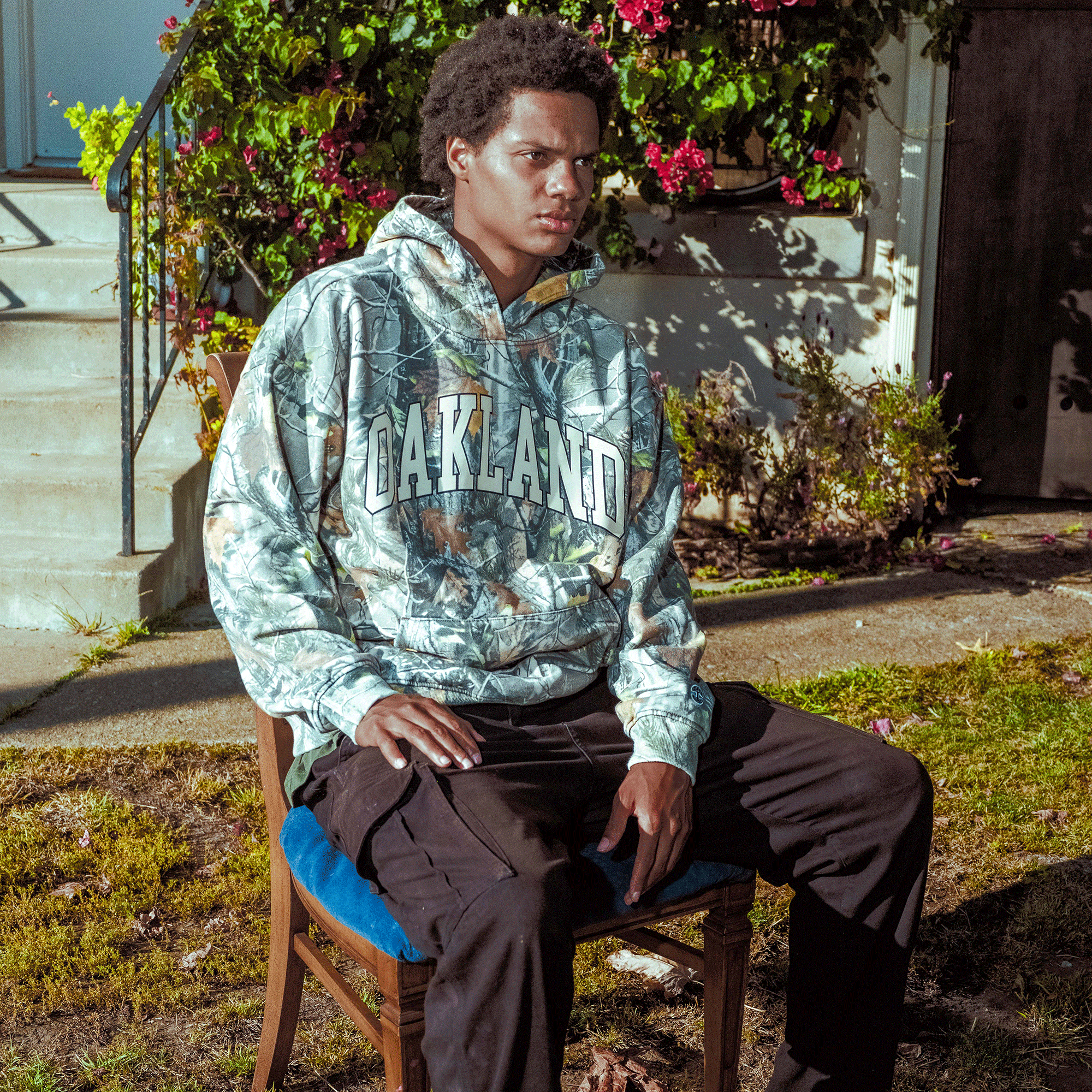 A person with short curly hair sits outdoors on a wooden chair with a blue cushion. They wear the Oaklandish Foliage Hoodie and black pants. Behind them are steps, green bushes, and vibrant pink flowers in sunlight.