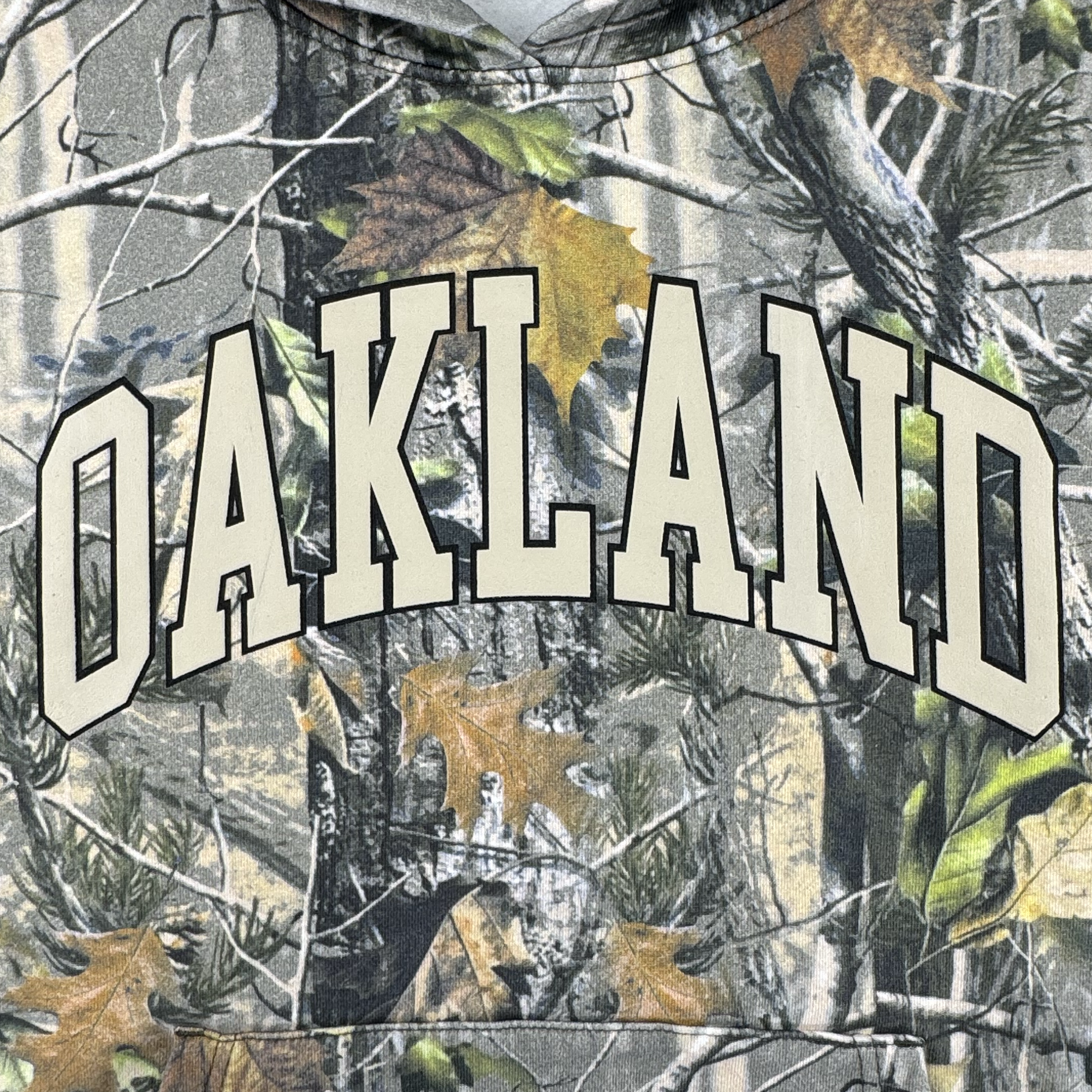 The Oaklandish Foliage Hoodie features a wood camo forest print with branches and autumn leaves, "OAKLAND" in bold cream letters with a black outline across the chest, and an embroidered Oaklandish tree detail.