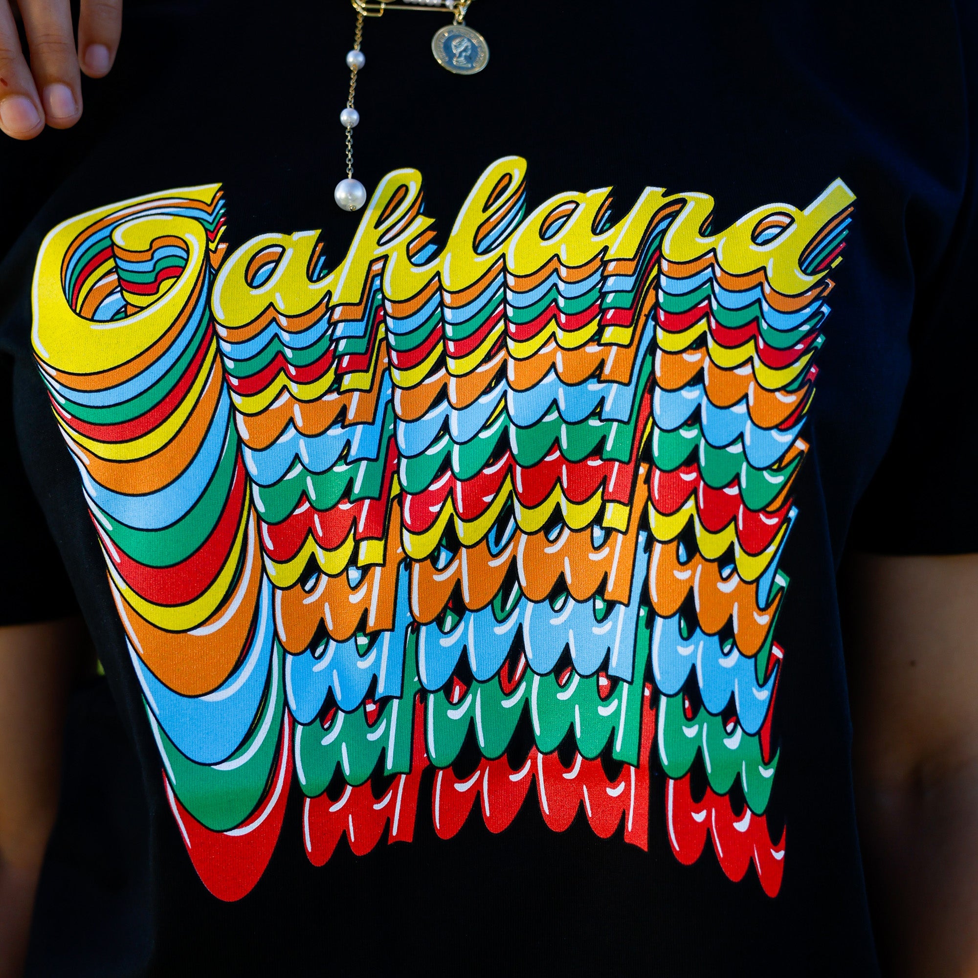 Close-up of a black Oakland Roots SC Funk Tee showcasing a vibrant, retro-style graphic design with the words "Oakland Roots" repeated multiple times. The design features colorful, layered letters in yellow, red, blue, and green. The person wearing the tee accessorizes with a necklace that has a pendant and a pearl chain.